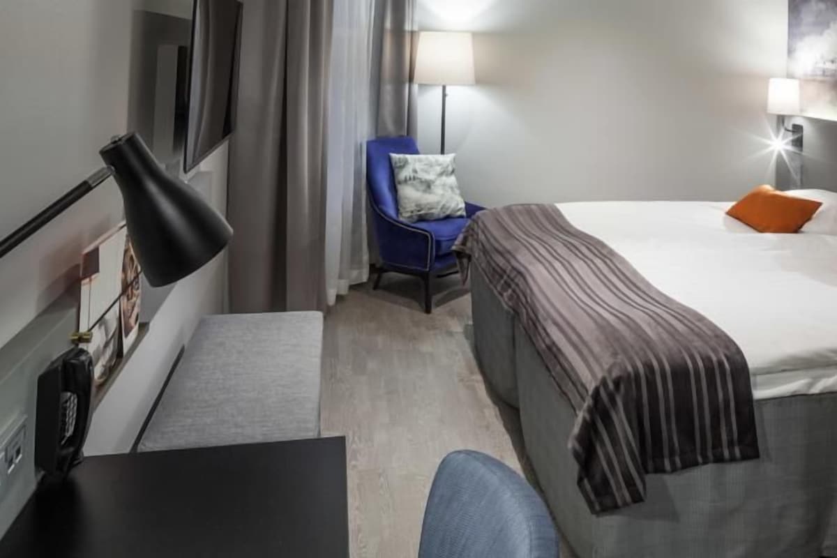 A hotel room with a bed, a desk, a chair, and a lamp.