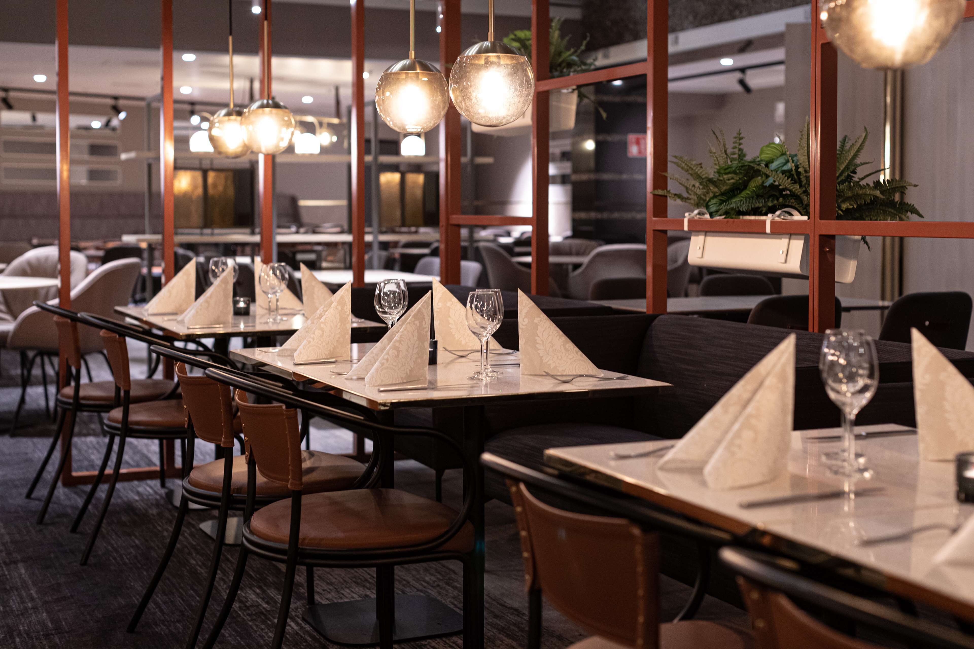 A restaurant with tables set for dining, featuring a modern interior with pendant lighting and a view of the cityscape through large windows.