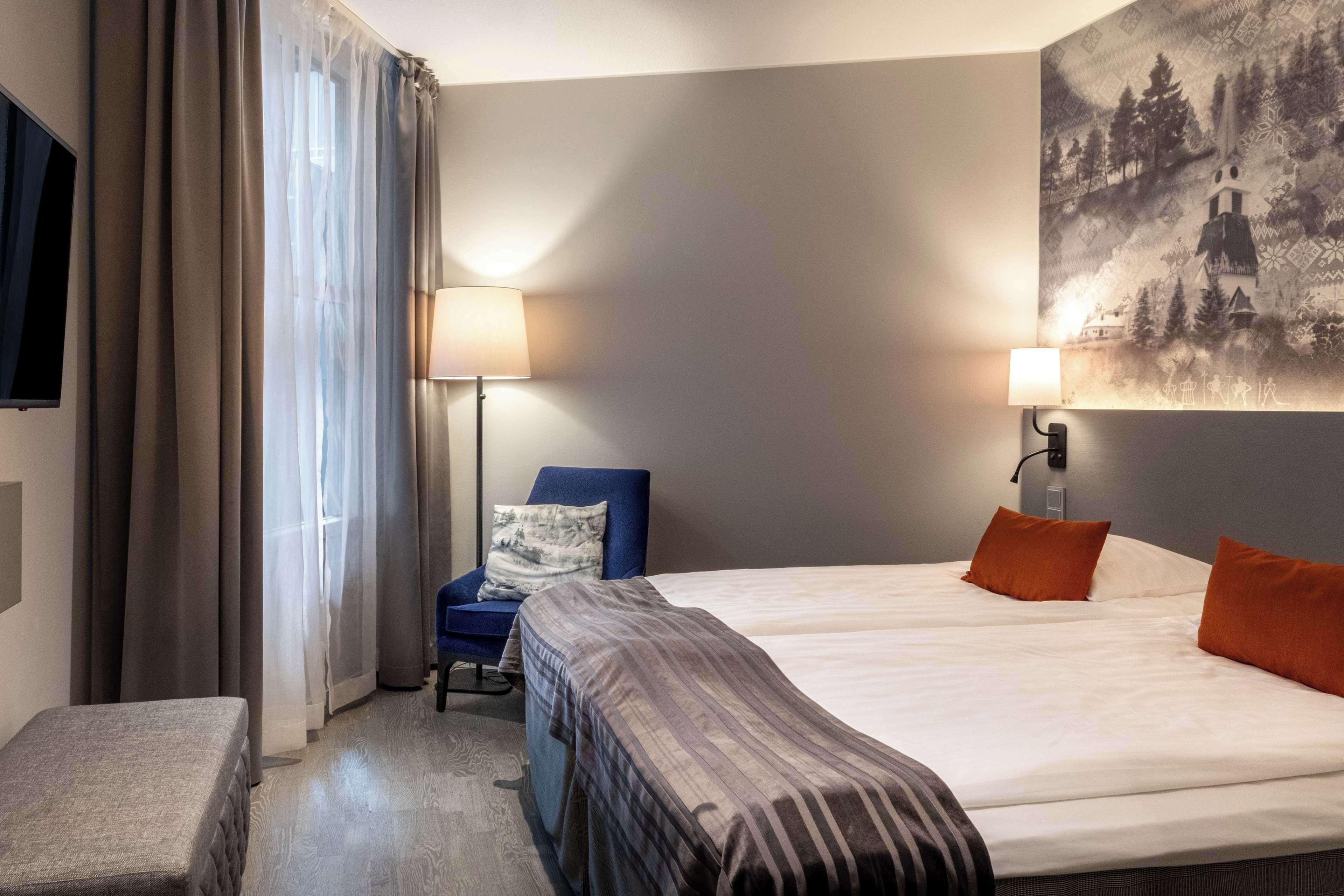 A hotel room with a bed, a chair, a desk, a lamp, and a painting on the wall.