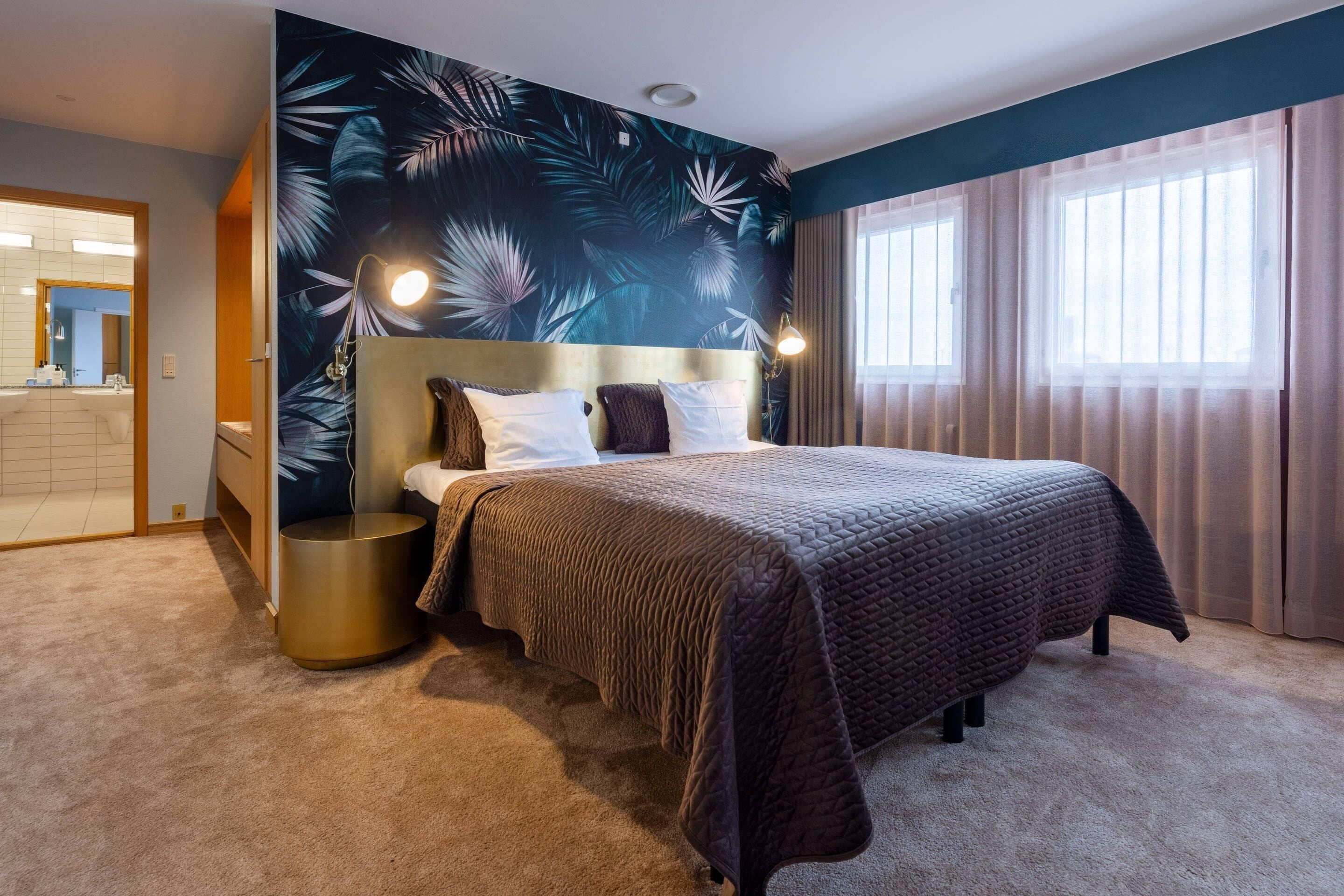 Une chambre à coucher avec un grand lit, une tête de lit, une table de chevet ronde et un mur orné d’un motif de feuilles tropicales.