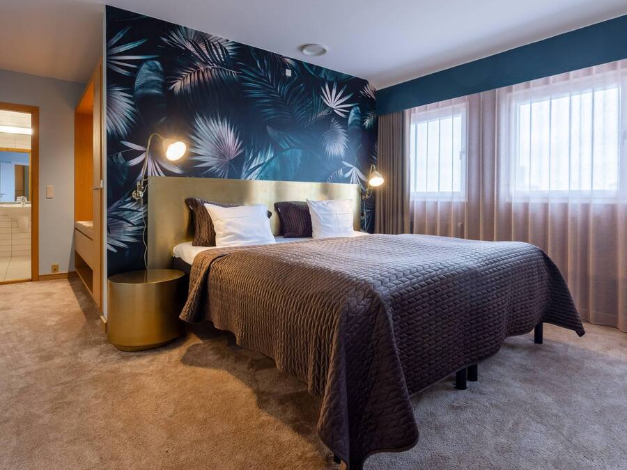 Une chambre à coucher avec un grand lit, une tête de lit, une table de chevet ronde et un mur orné d’un motif de feuilles tropicales.