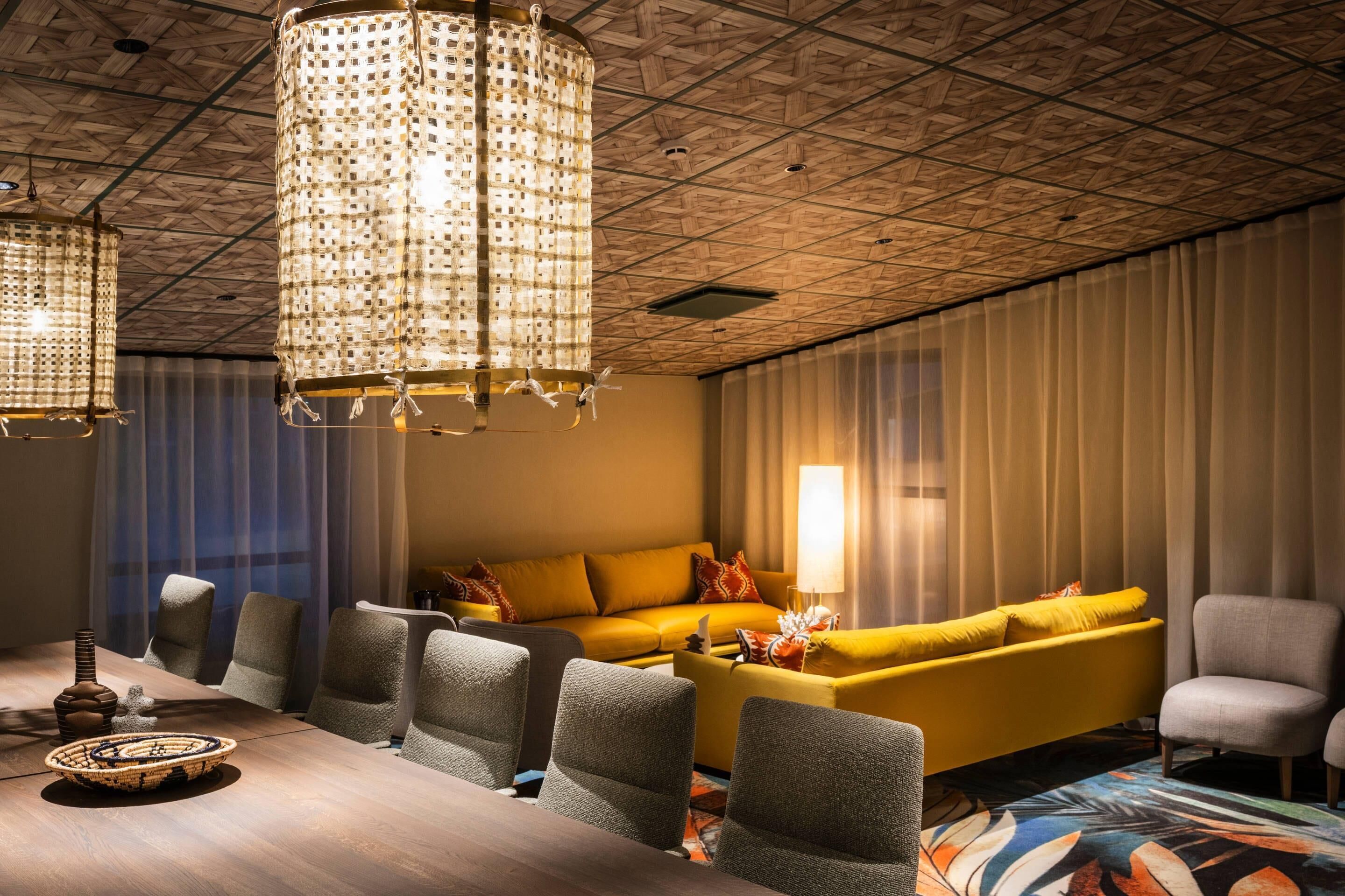 Una habitación de hotel moderna con techo de madera, un sofá amarillo, sillones grises, una mesa de comedor con sillas y una lámpara de araña con motivos decorativos.