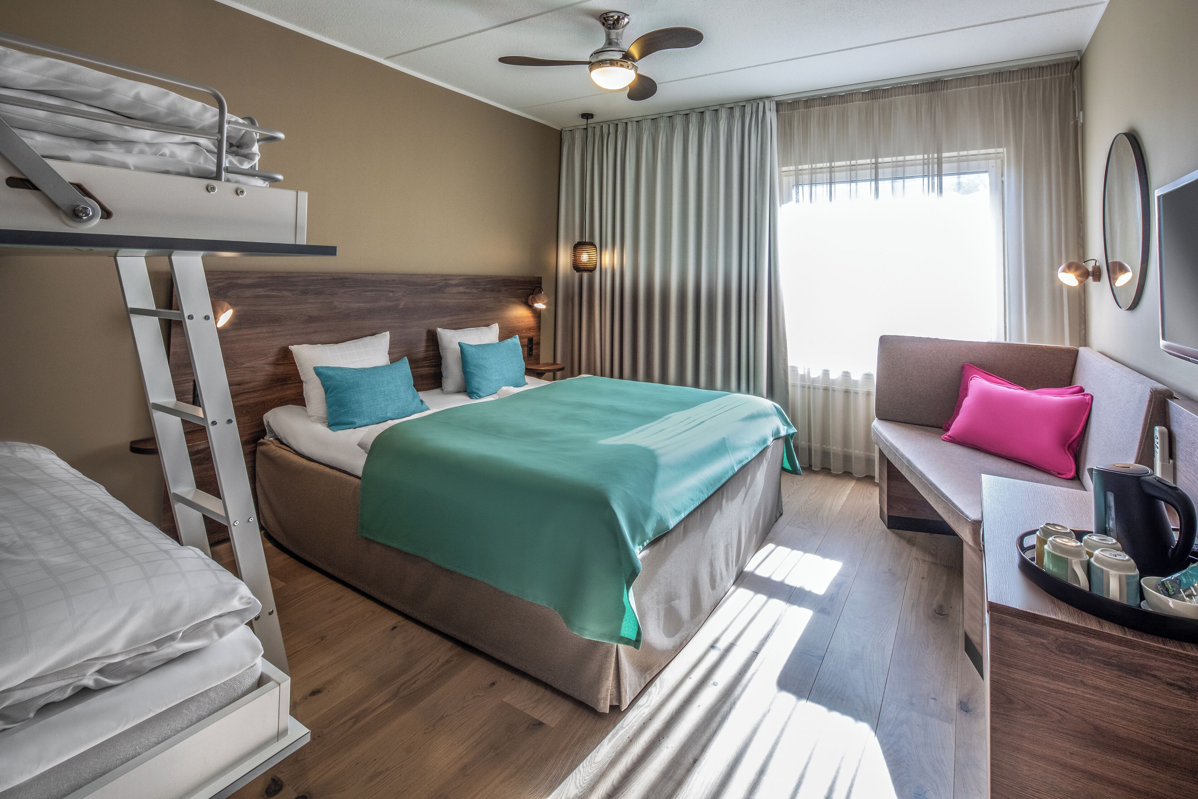Habitación de hotel con literas, un sofá, un escritorio con una bandeja de objetos y un ventanal con cortinas.