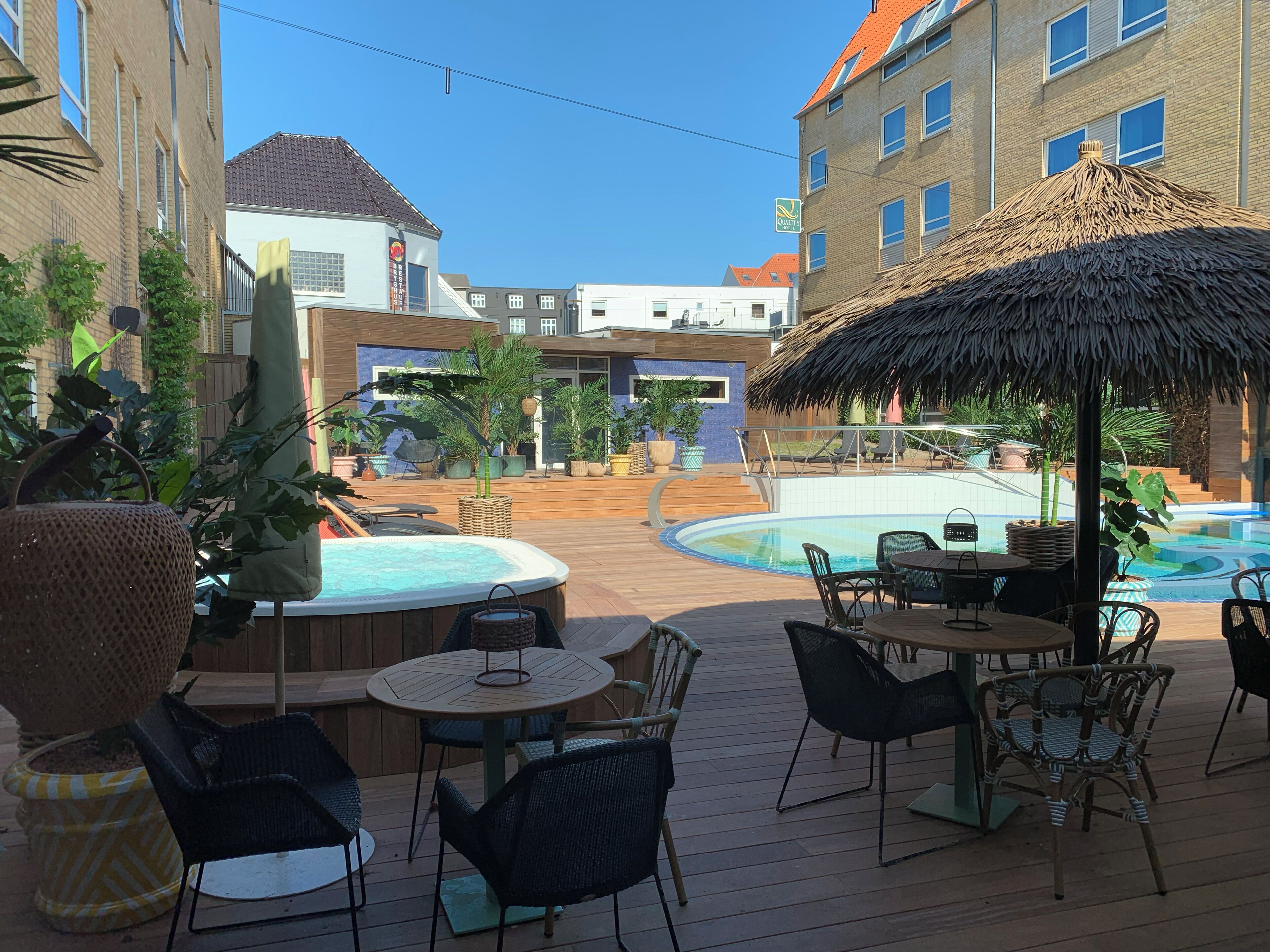 A rooftop pool area with lounge chairs and a thatched umbrella, surrounded by potted plants and a view of the cityscape.