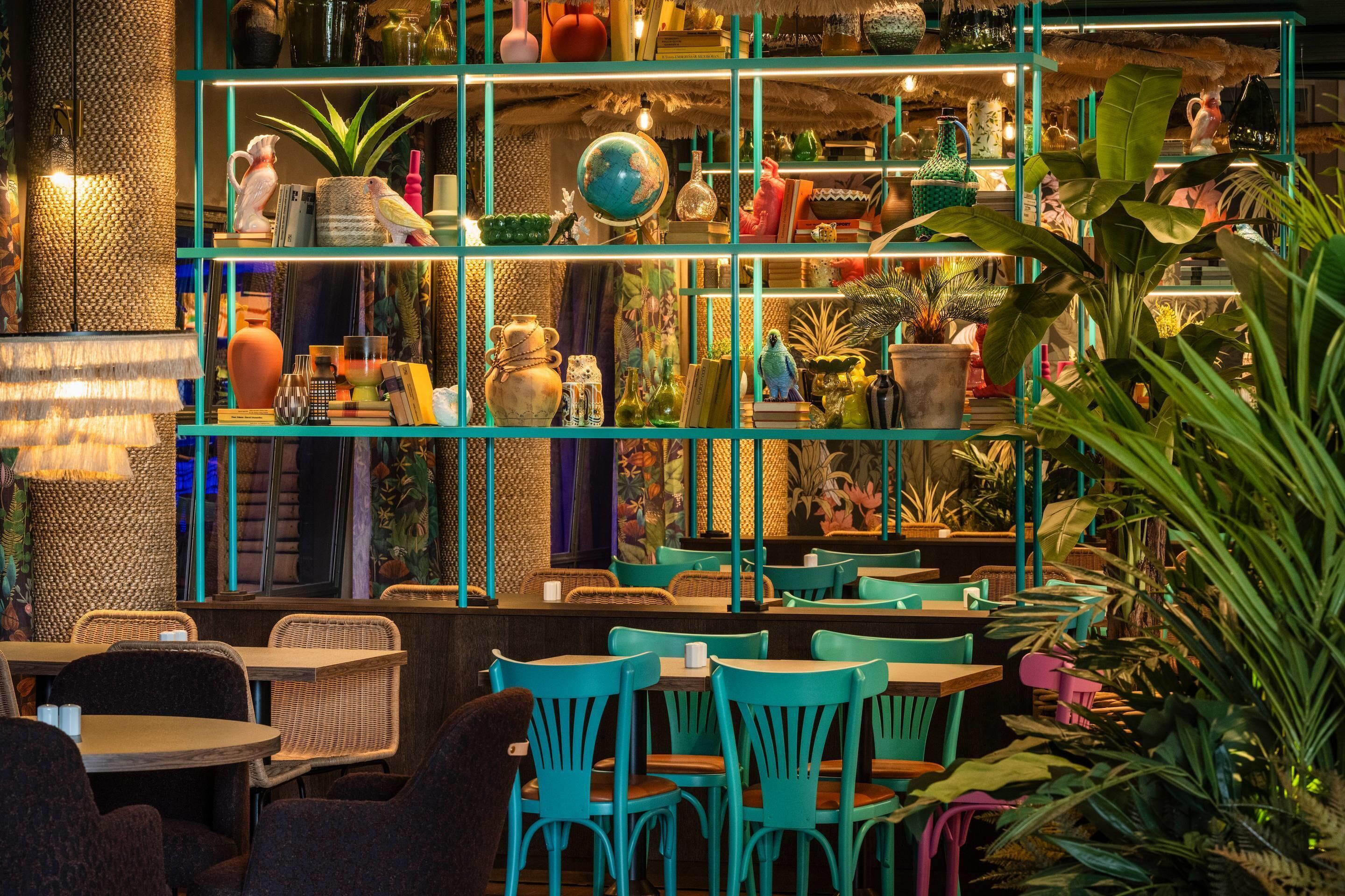 Un restaurant avec des chaises et des tables de couleur turquoise, des étagères remplies de divers objets et une décoration d’inspiration tropicale.