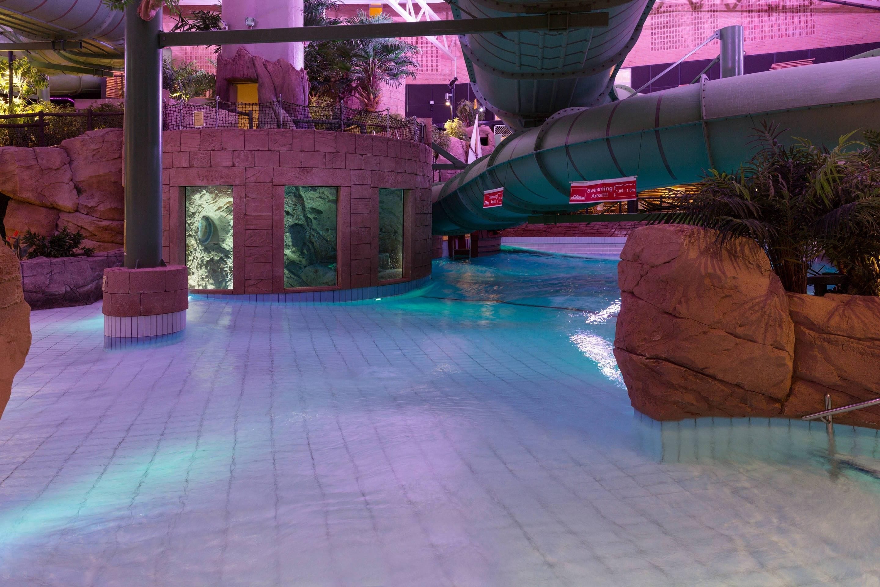 Parque acuático interior con una piscina grande, formaciones rocosas y un tobogán.