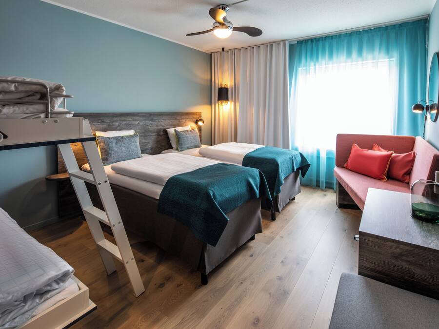 Habitación de hotel con dos camas, un escritorio, una silla, un ventilador de techo y una ventana con cortinas.