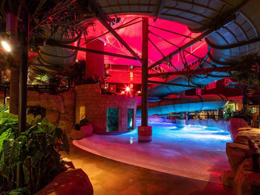 Parque aquático interno com uma piscina grande, plantas tropicais e teto iluminado em vermelho.