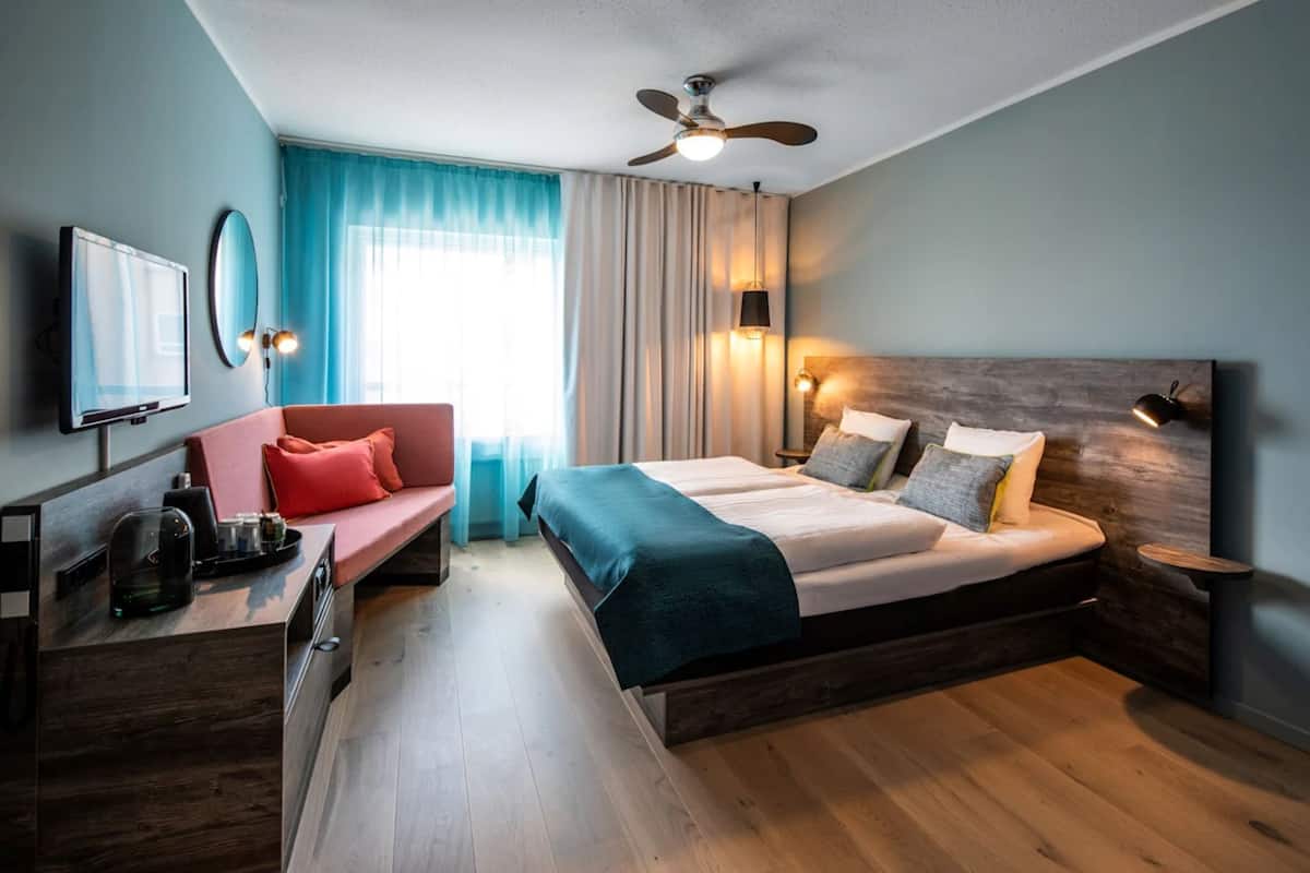 A hotel room with a bed, a desk, a chair, a ceiling fan, and a TV.
