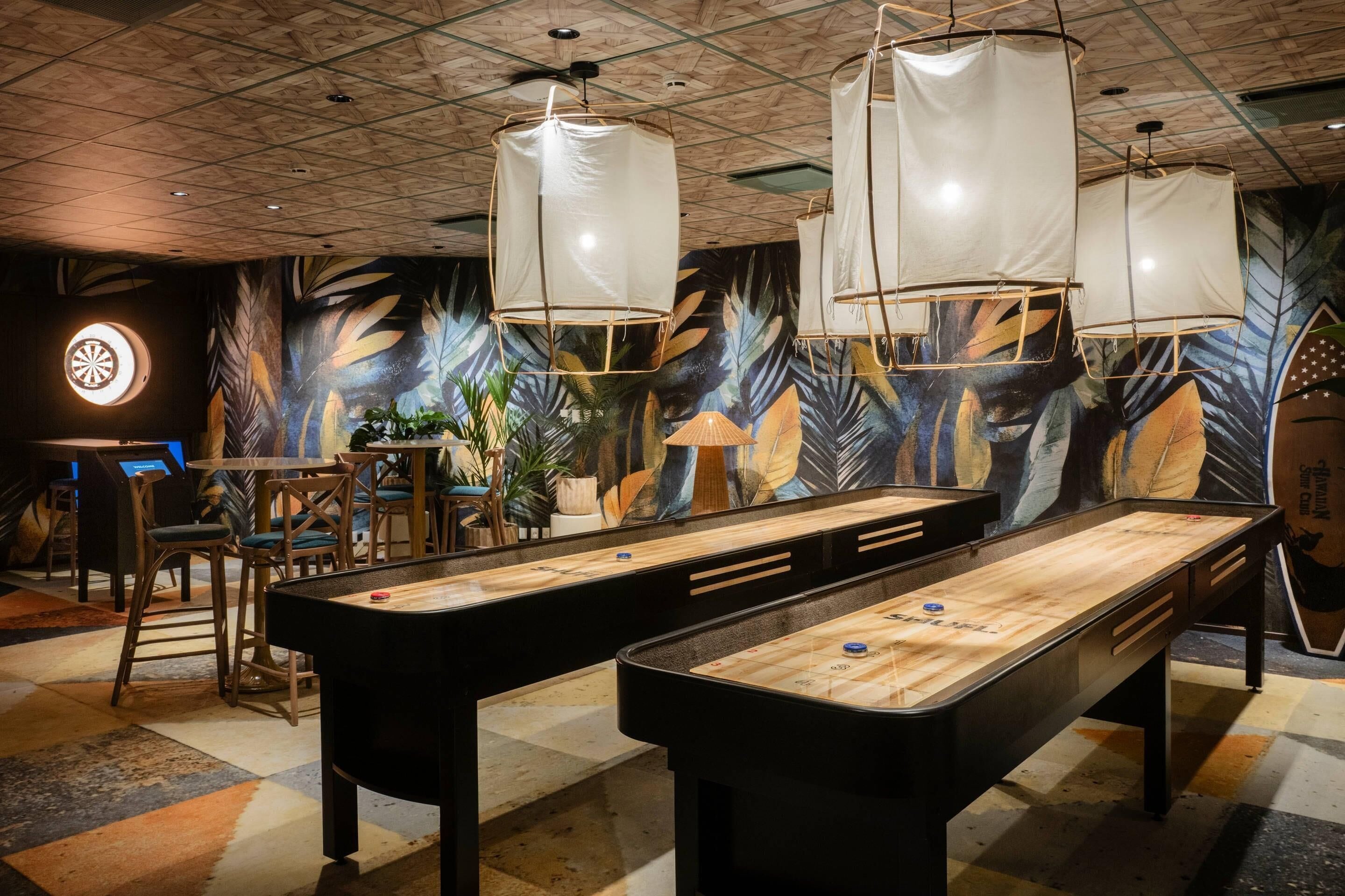 Une table de billard dans une pièce dont un mur est décoré d’une fresque représentant des feuilles tropicales, avec des chaises en bois et des lampes suspendues.