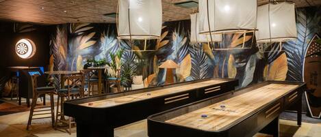 Une table de billard dans une pièce dont un mur est décoré d’une fresque représentant des feuilles tropicales, avec des chaises en bois et des lampes suspendues.