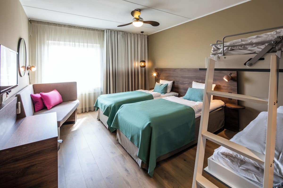 A hotel room with two beds, a desk, a bunk bed, a ceiling fan, and a window with curtains.