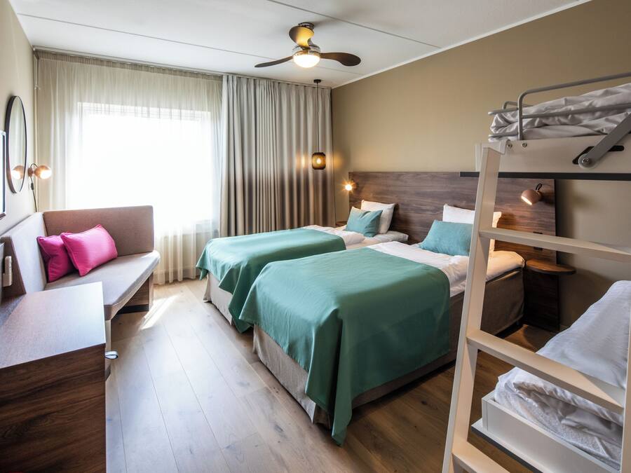 A hotel room with two beds, a desk, a bunk bed, a ceiling fan, and a window with curtains.