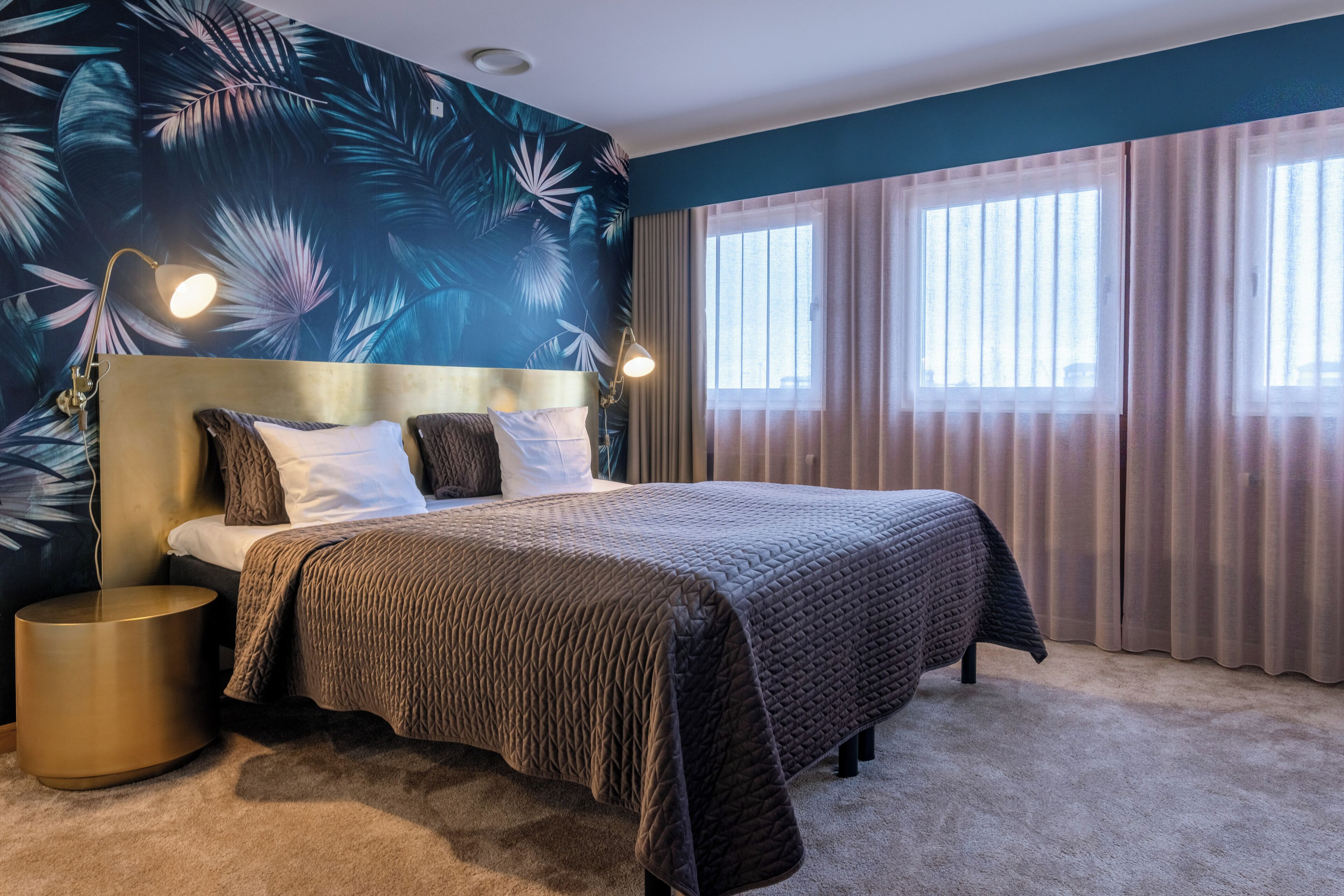 Un dormitorio con una cama grande, un cabecero, dos lámparas de noche, una mesita redonda y un papel tapiz con estampado de hojas tropicales.