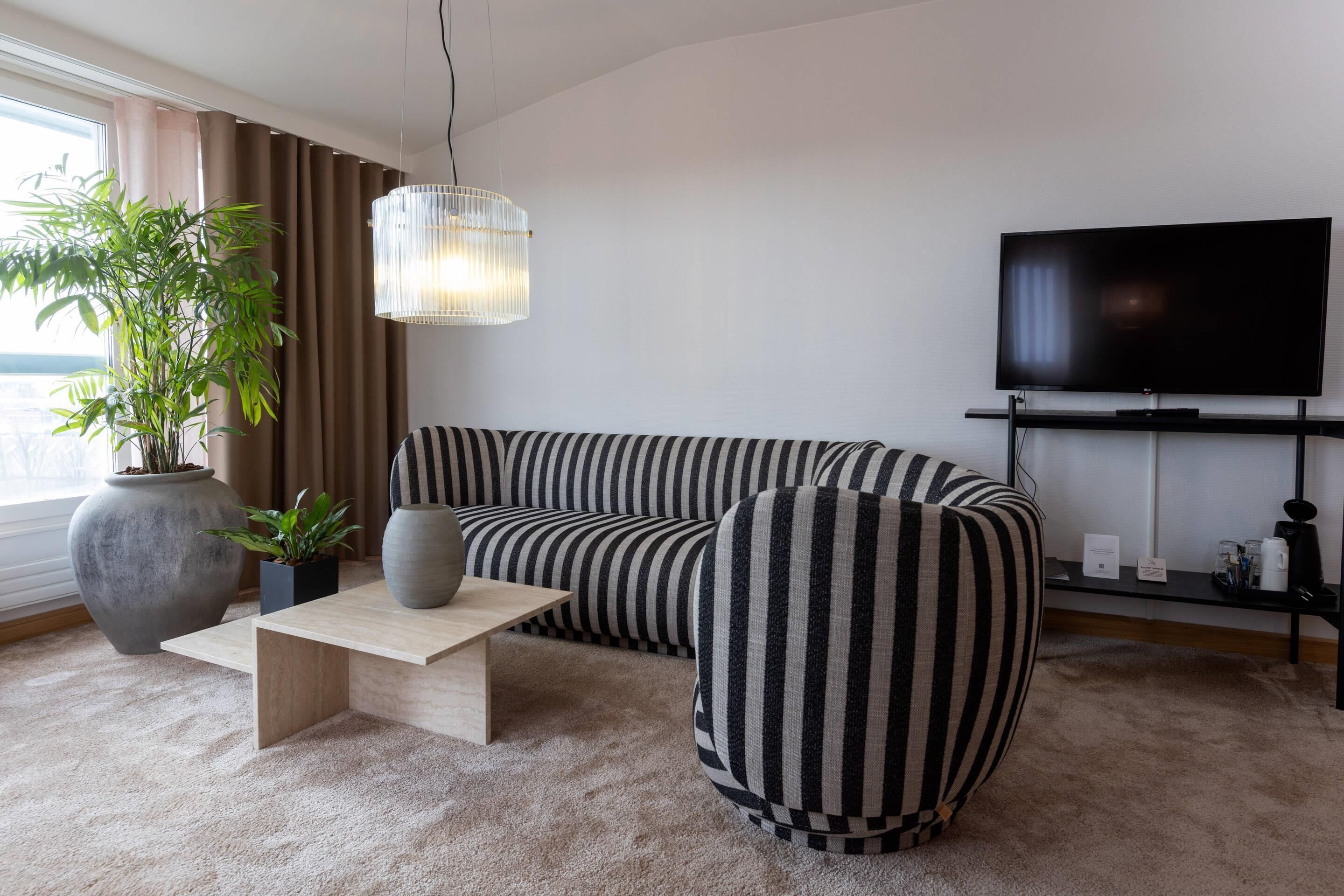 A modern living room with a striped sofa, a wooden coffee table, a large potted plant, and a flat-screen TV on a stand.