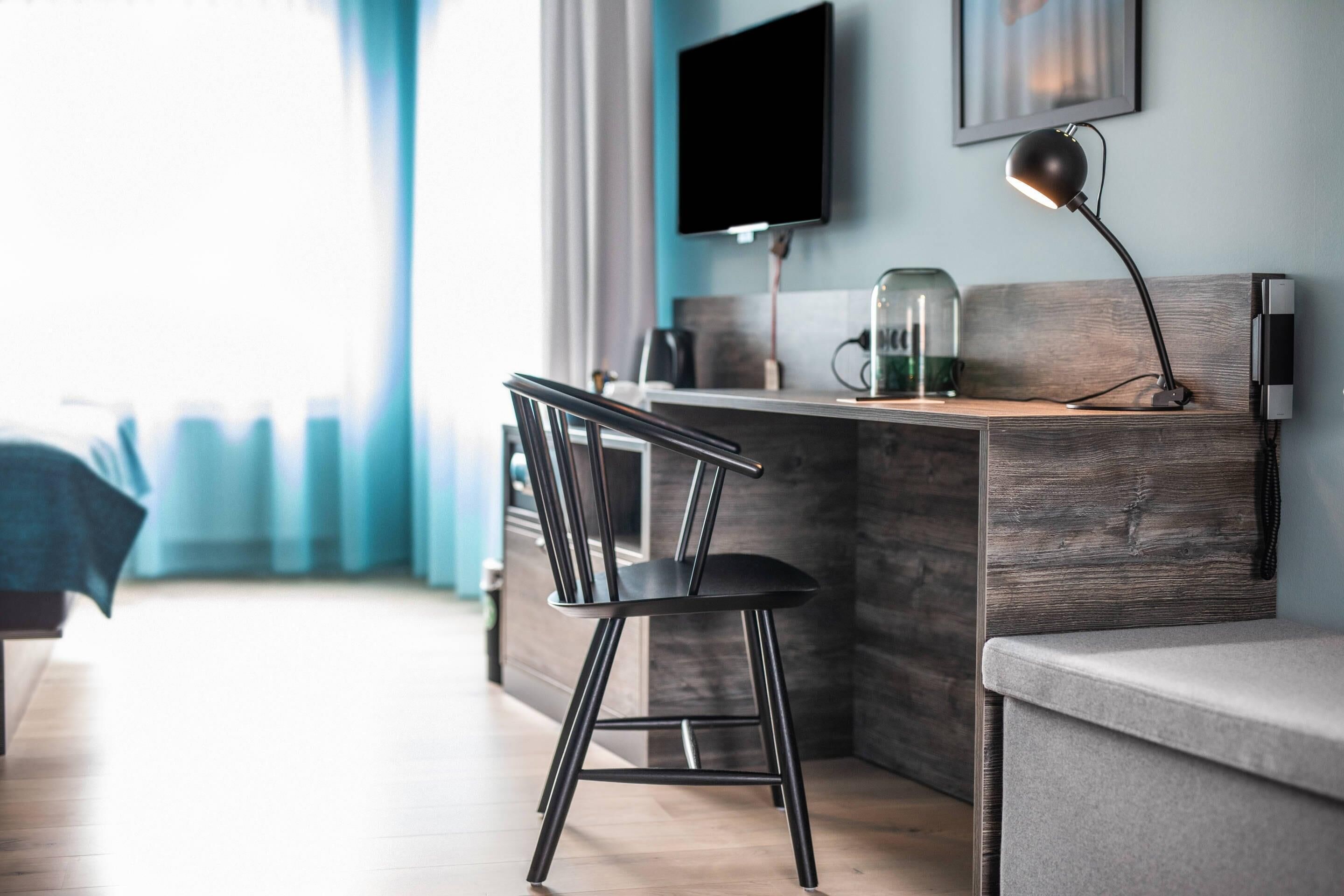 Een moderne hotelkamer met een houten bureau, een zwarte stoel, een flatscreen televisie aan de muur en een nachtkastje met een lamp.