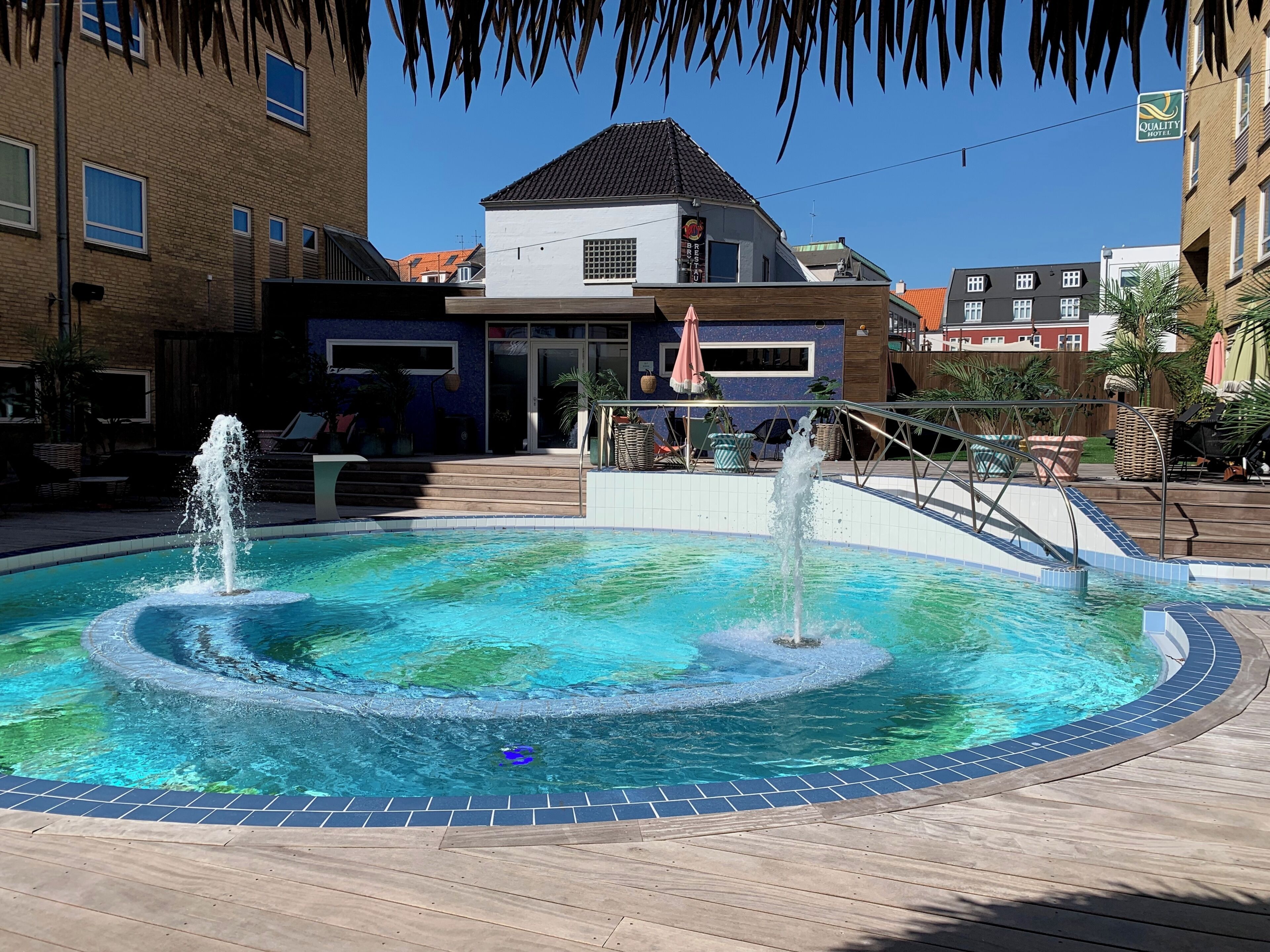 A swimming pool with two fountains, surrounded by a wooden deck and a building with a sign that reads 'Quality Hotel.'
