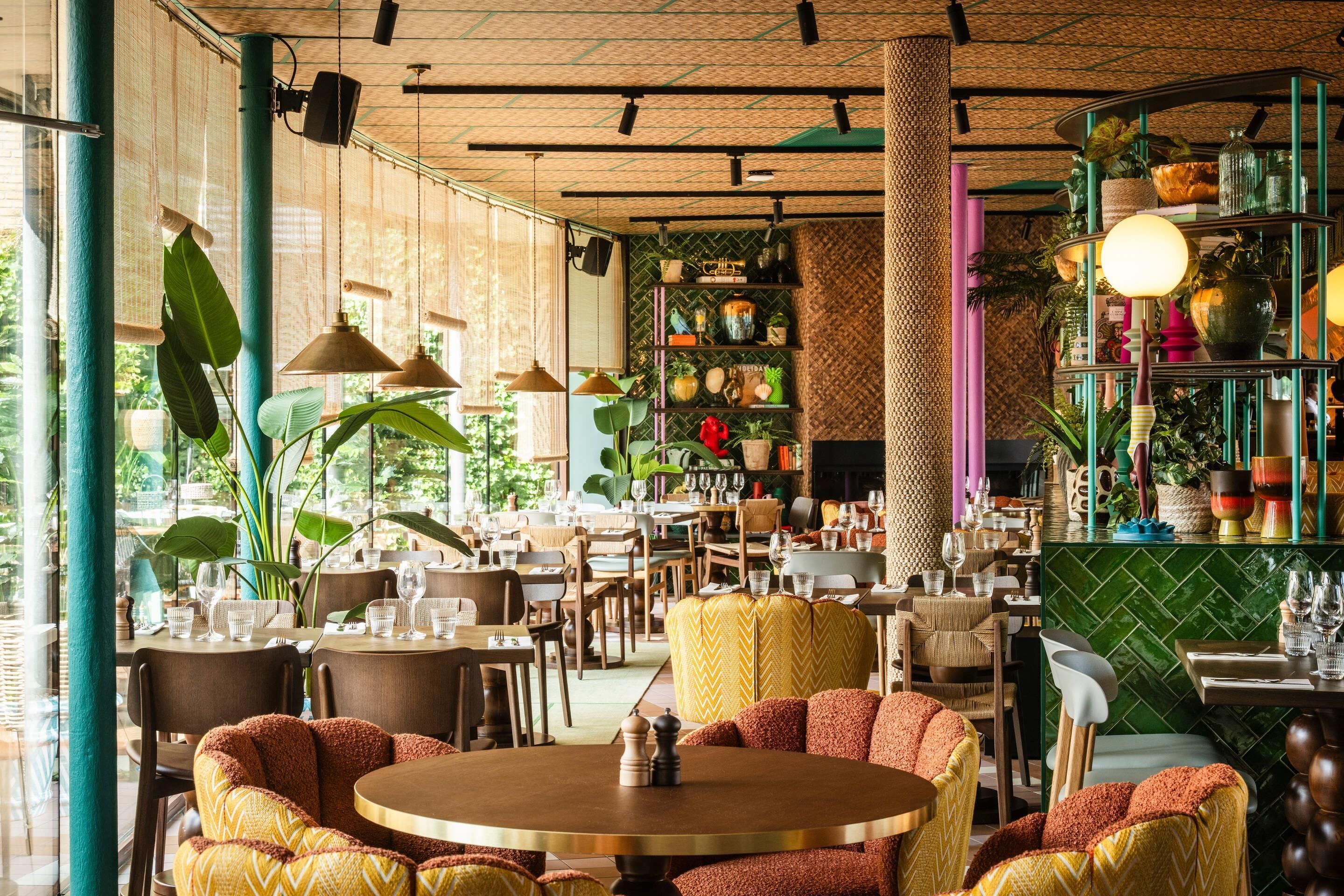 L’intérieur d’un restaurant, avec des tables rondes, des chaises, des luminaires suspendus et divers objets décoratifs disposés sur des étagères.