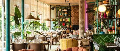 L’intérieur d’un restaurant, avec des tables rondes, des chaises, des luminaires suspendus et divers objets décoratifs disposés sur des étagères.