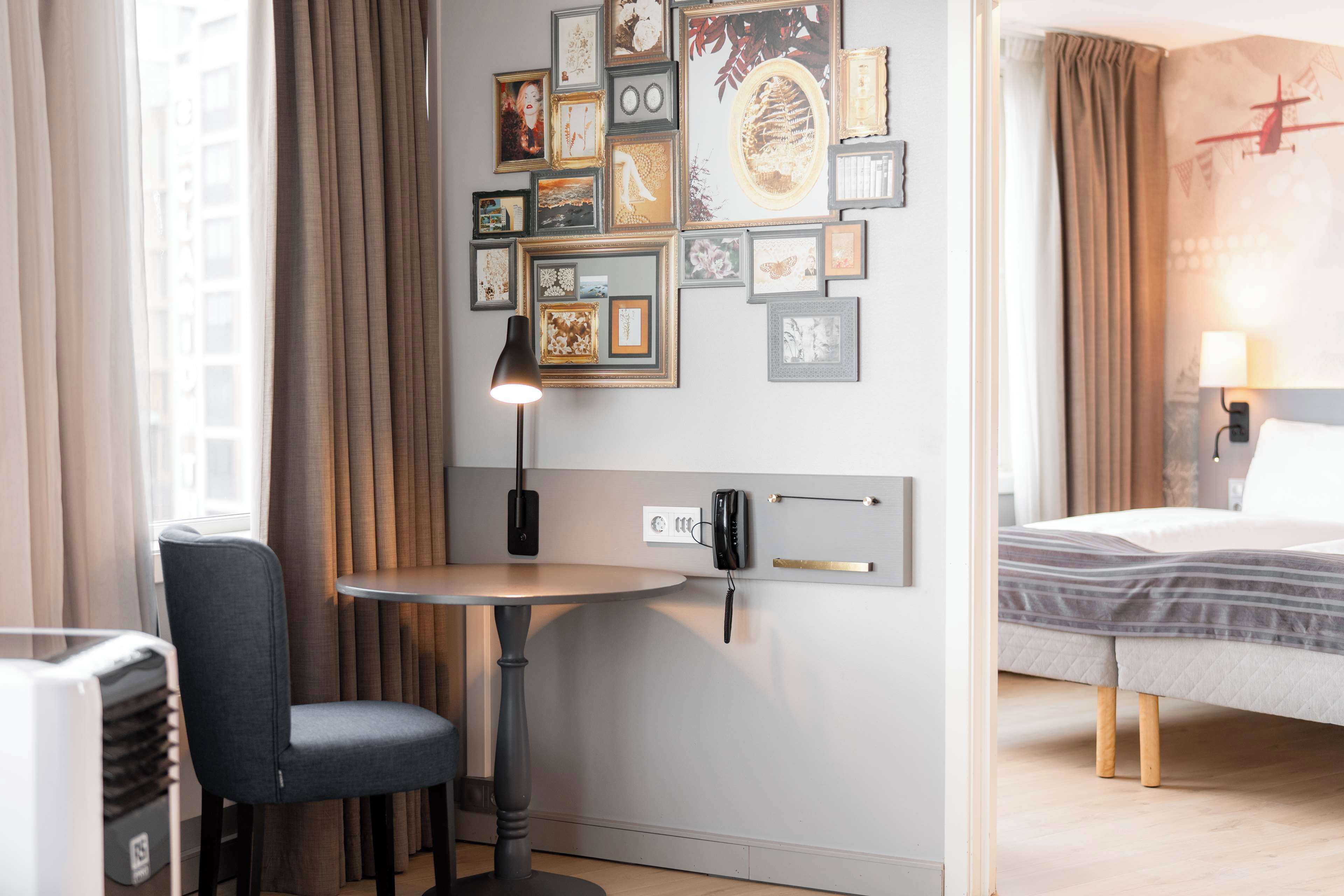 Quarto de hotel com uma cama, uma escrivaninha com cadeira, uma mesinha e um painel decorado com quadros.