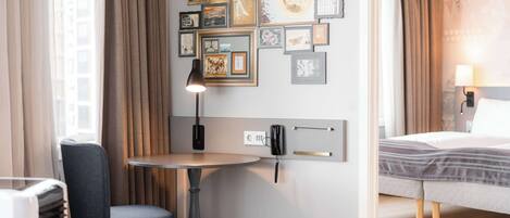 A hotel room with a bed, a desk with a chair, a small table, and a wall decorated with framed pictures.
