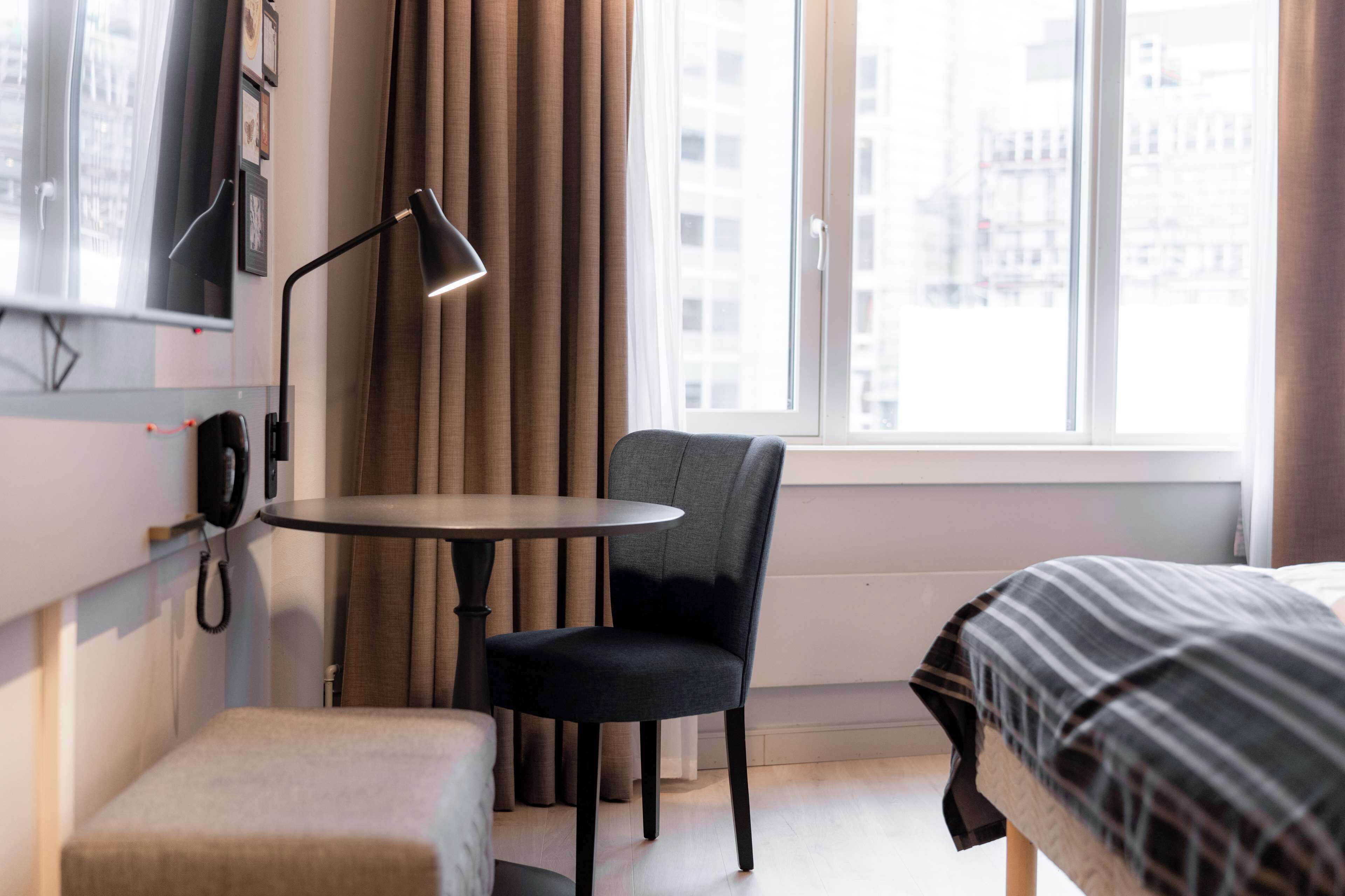 A hotel room with a bed, a small table, a chair, a desk lamp, a telephone, and a window with curtains.