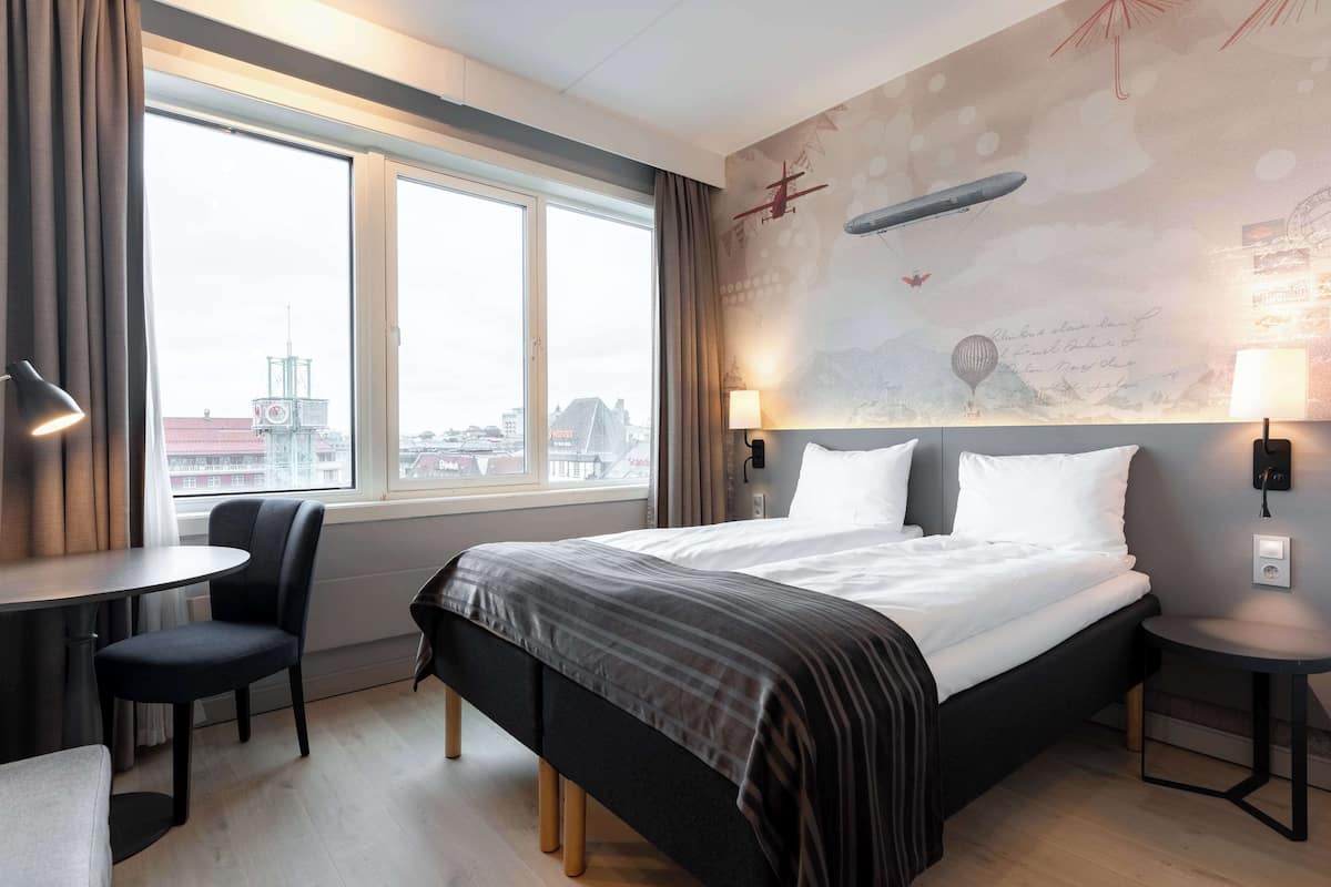 A hotel room with a large bed, two bedside tables, a chair, and a view of the city through a large window.