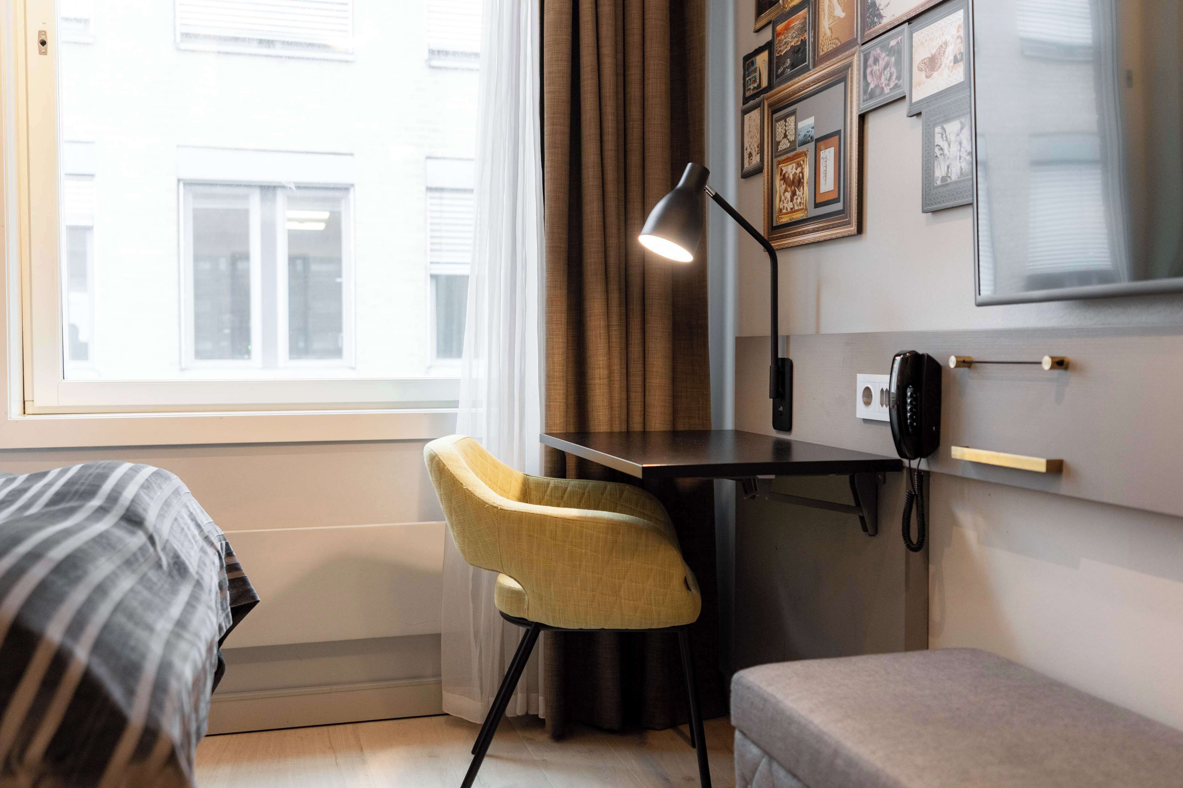 Ett hotellrum med en säng, ett skrivbord med en lampa, en stol, en telefon och ett fönster med gardiner.