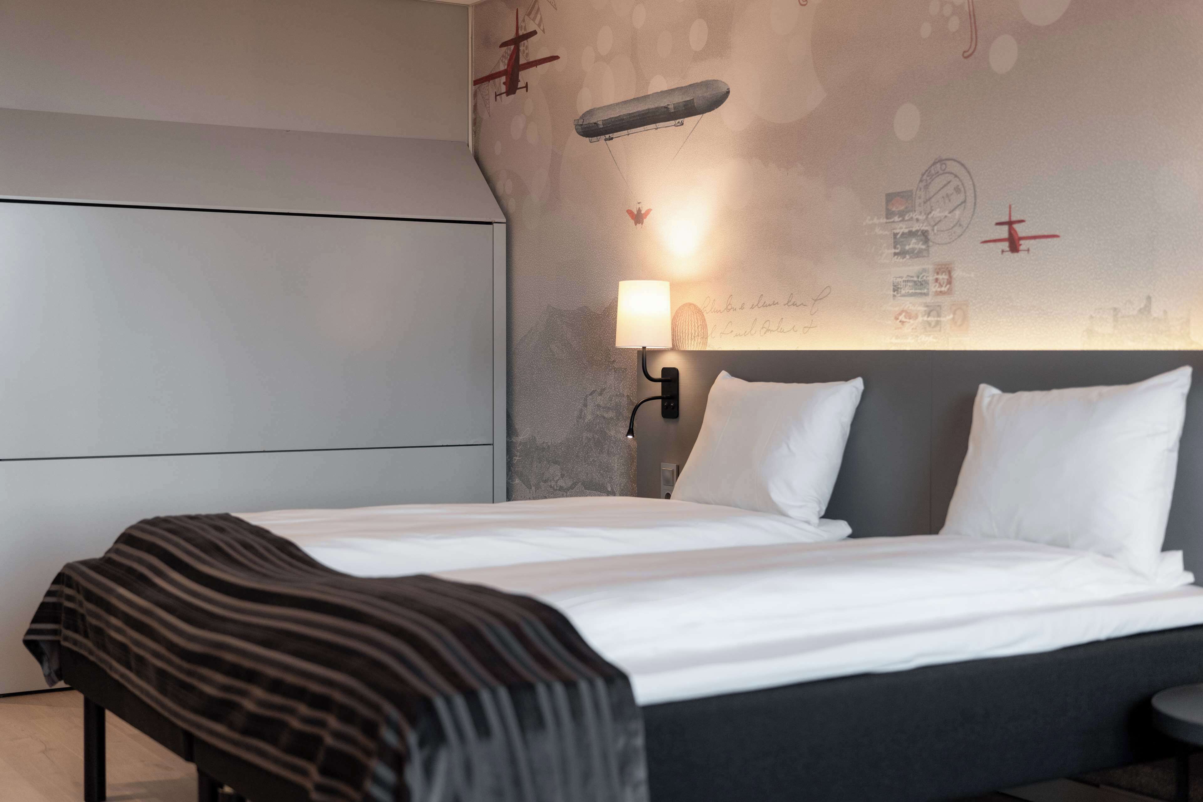 Una habitación de hotel moderna con una cama grande, una mesita de noche con lámpara y una pared decorada con adhesivos de aviones antiguos.