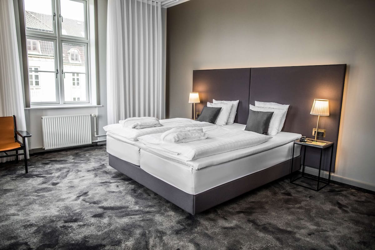 Moderni hotellihuone, jossa on suuri sänky, kaksi yöpöytävalaisinta, tuoli ja ikkunasta avautuva näkymä kaupunkiin.