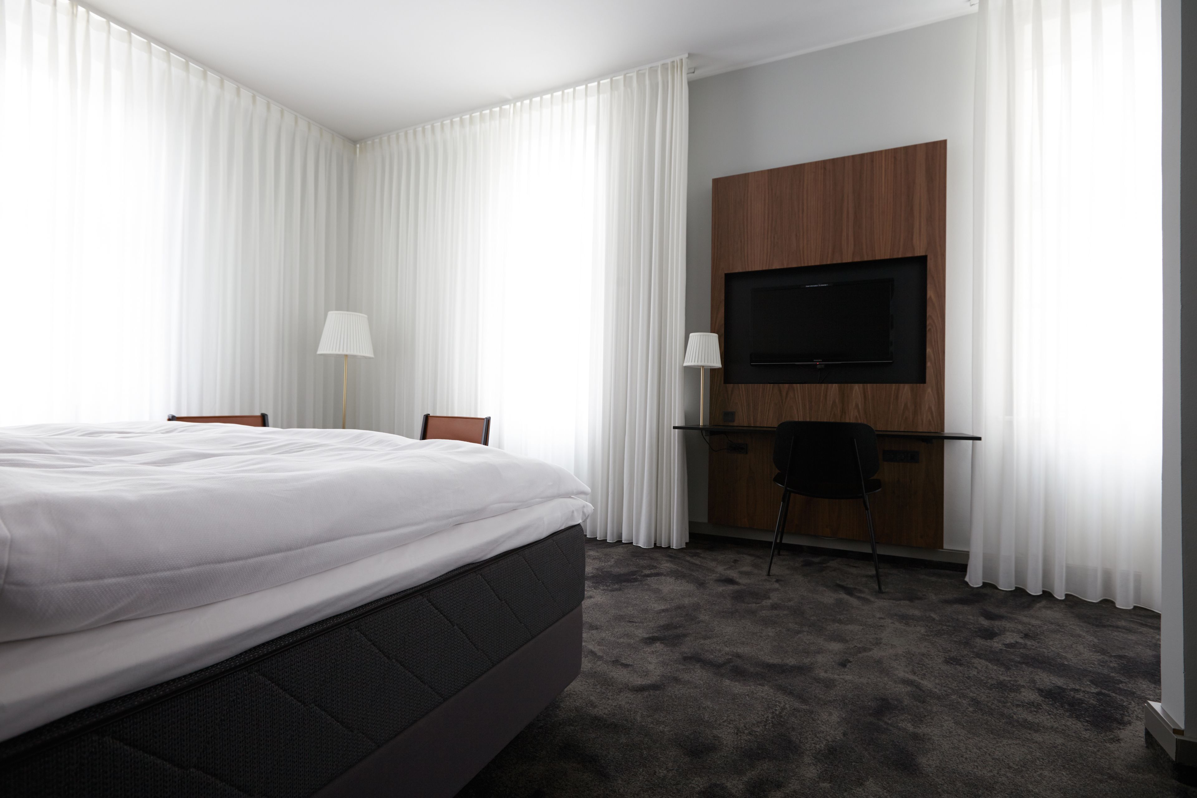 A hotel room with a large bed, a desk with a chair, a television mounted on a wooden panel, and sheer white curtains.