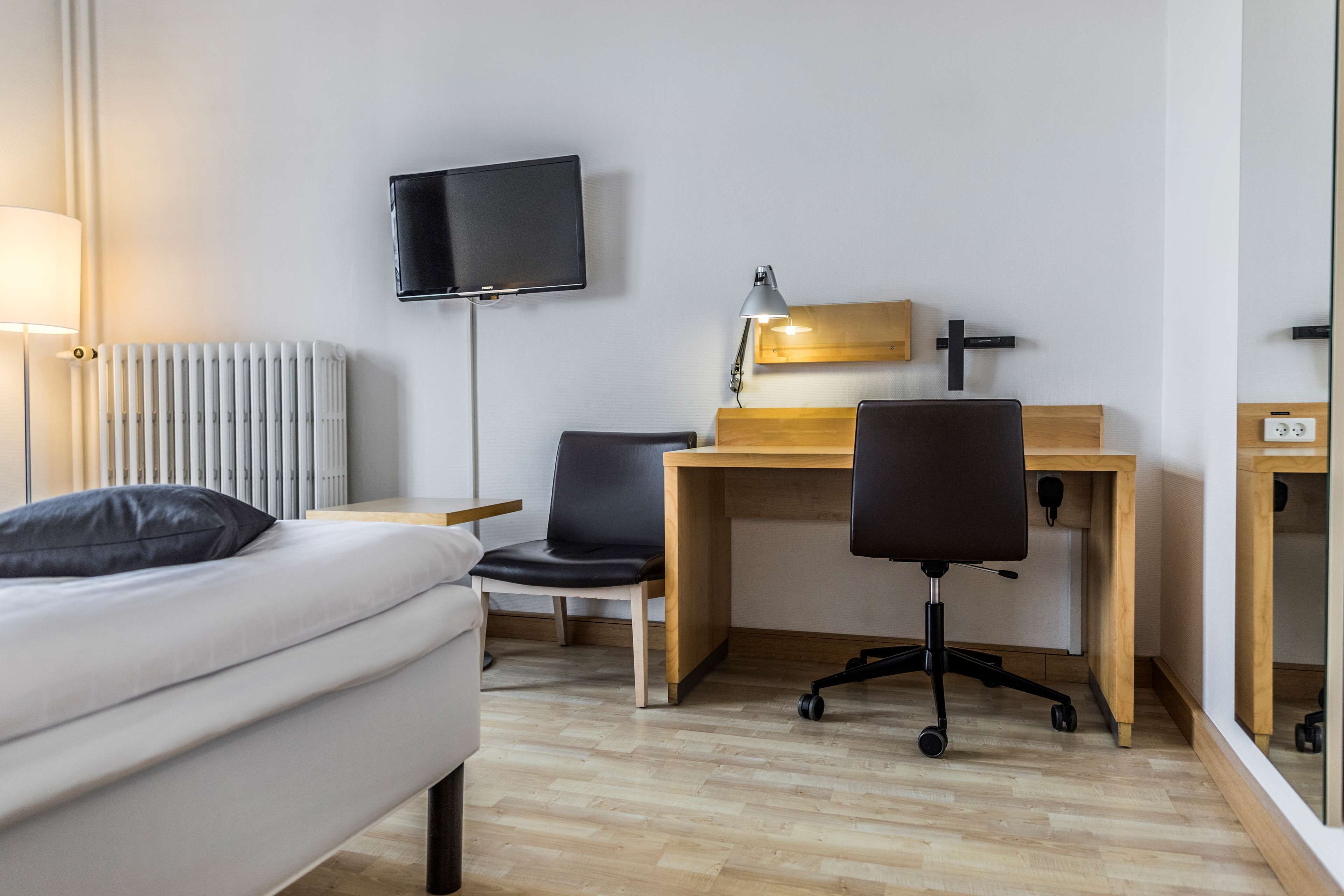 A hotel room with a bed, a desk, a chair, a TV, a lamp, and a radiator.