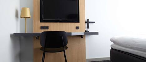 Habitación de hotel compacta con escritorio de madera, una silla negra, televisor montado en la pared, mesita de noche y cama con ropa blanca.