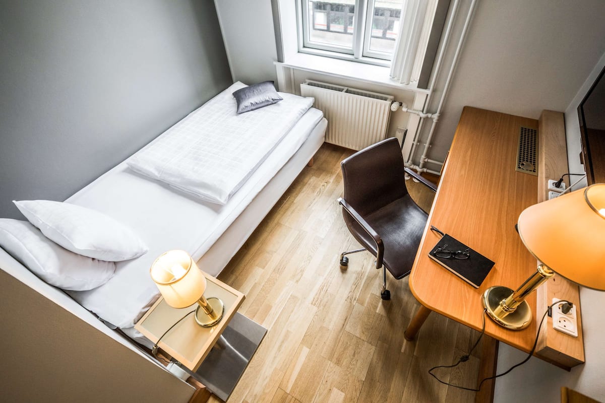 Pieni, siististi järjestetty huone, jossa on yksi sänky, työpöytä tuolilla, lamppu ja patteri.