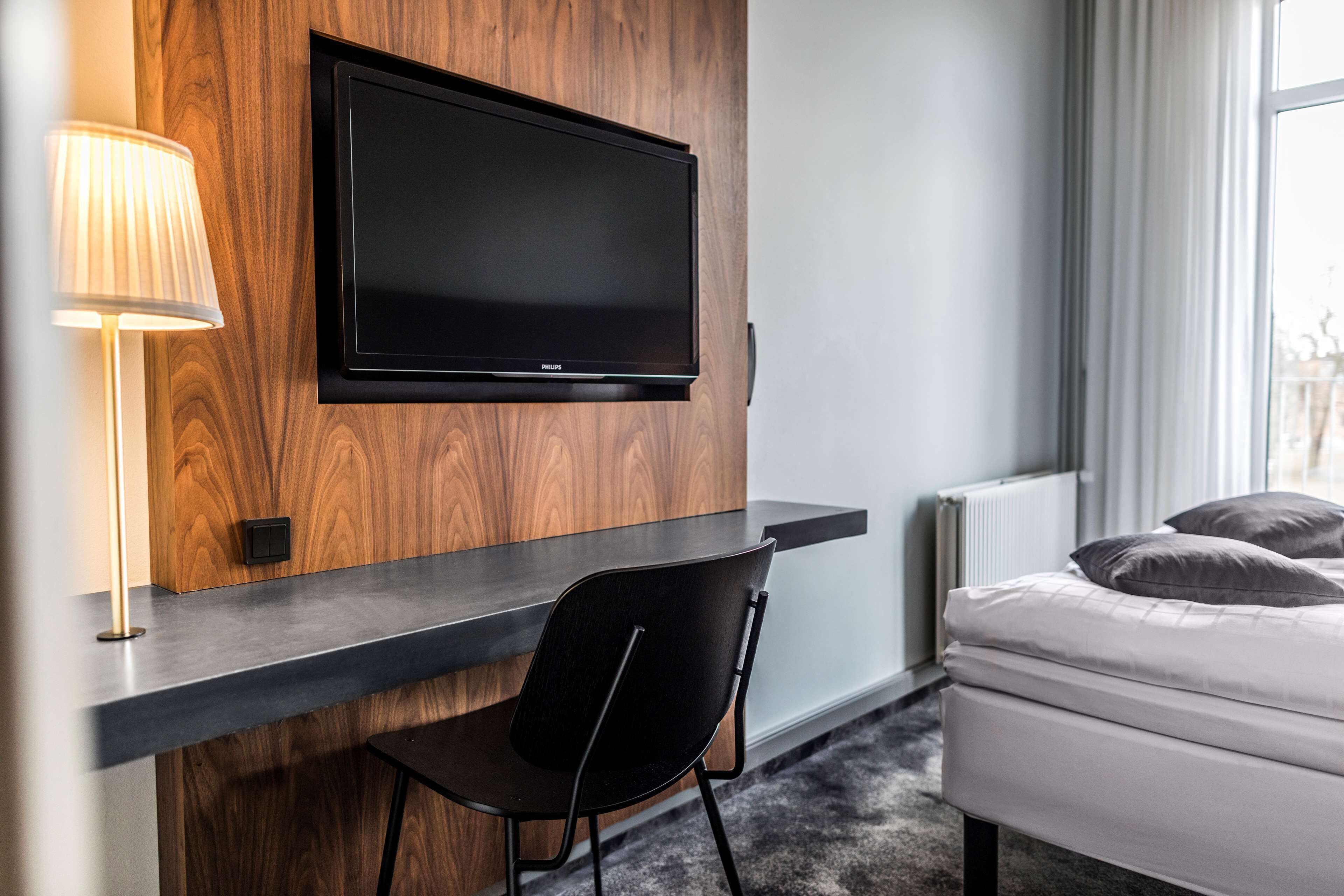 Ein modernes Hotelzimmer mit einem an der Wand befestigten Fernseher, einem Schreibtisch mit Stuhl, einem Bett mit Kissen und einer Lampe.