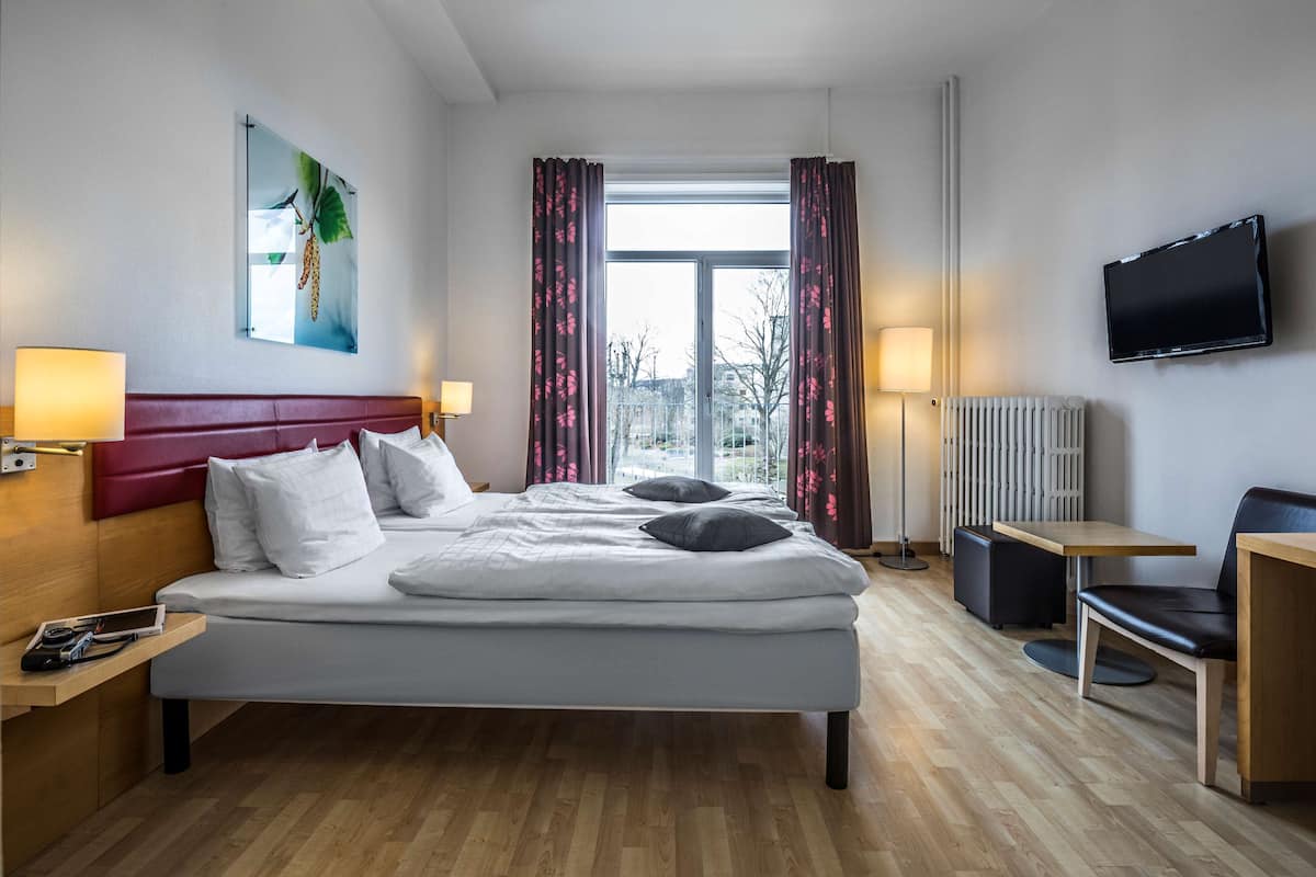 Hotellihuone, jossa on sänky, yöpöydät, työpöytä, tuoli, televisio, ikkuna verhoilla ja patteri.
