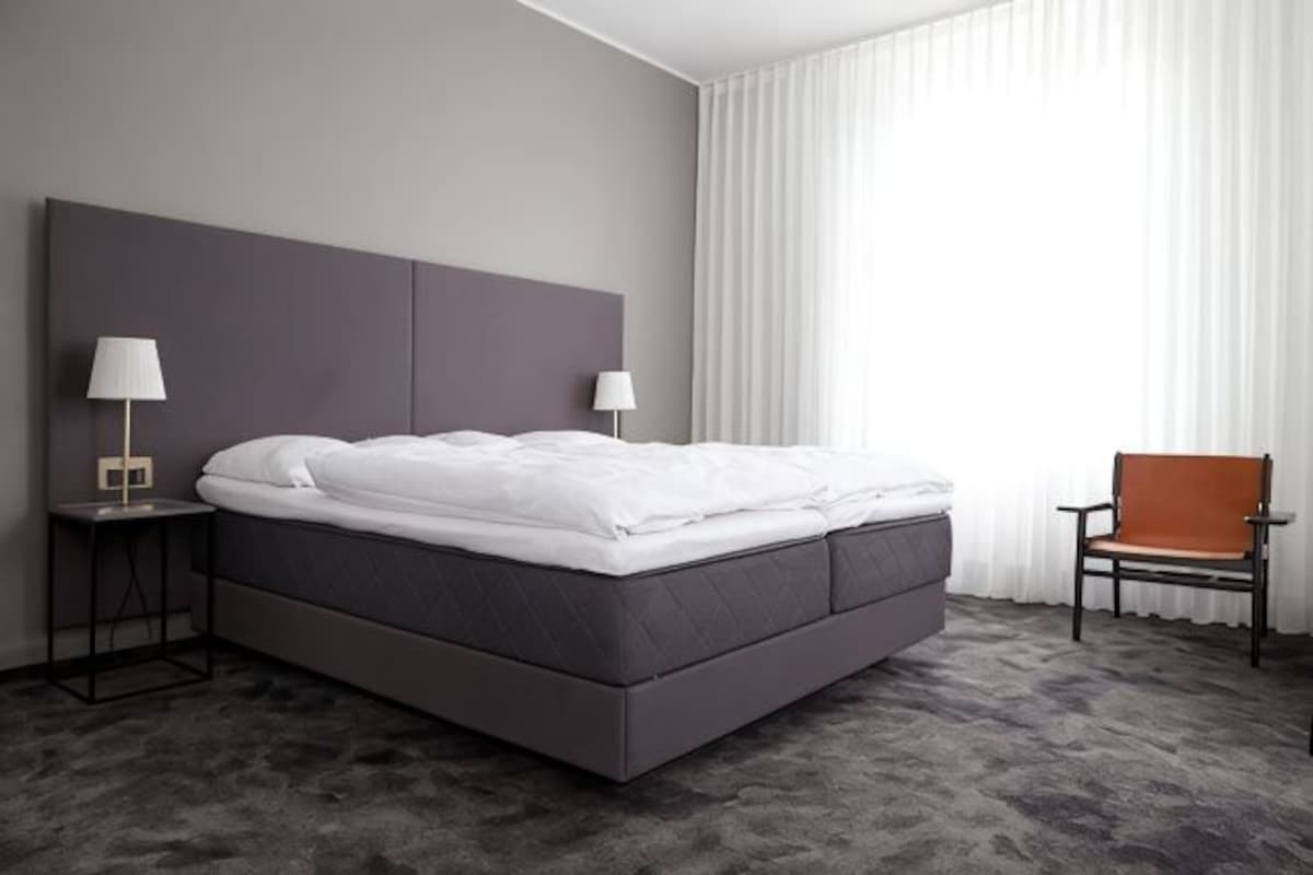 A modern hotel room with a large bed, two bedside tables with lamps, a chair, and a large window with curtains.