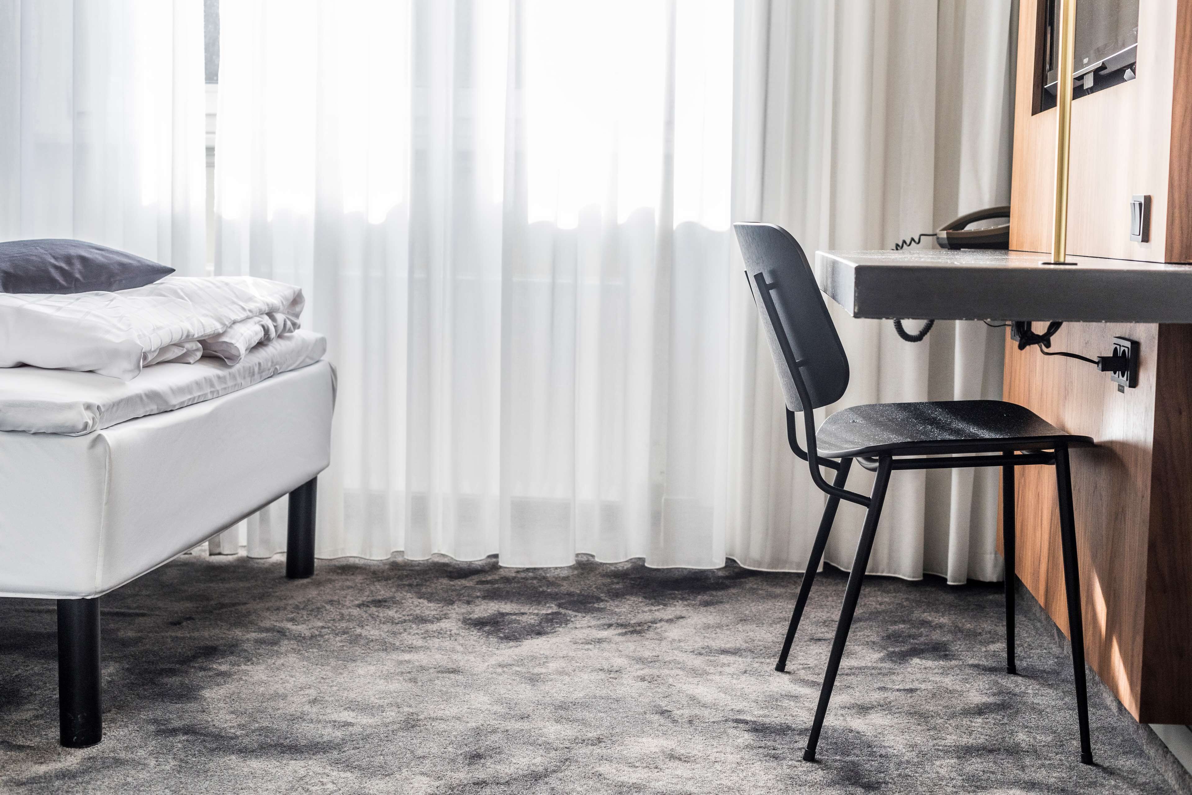 A hotel room with a bed, a desk, a chair, and a window with sheer curtains.