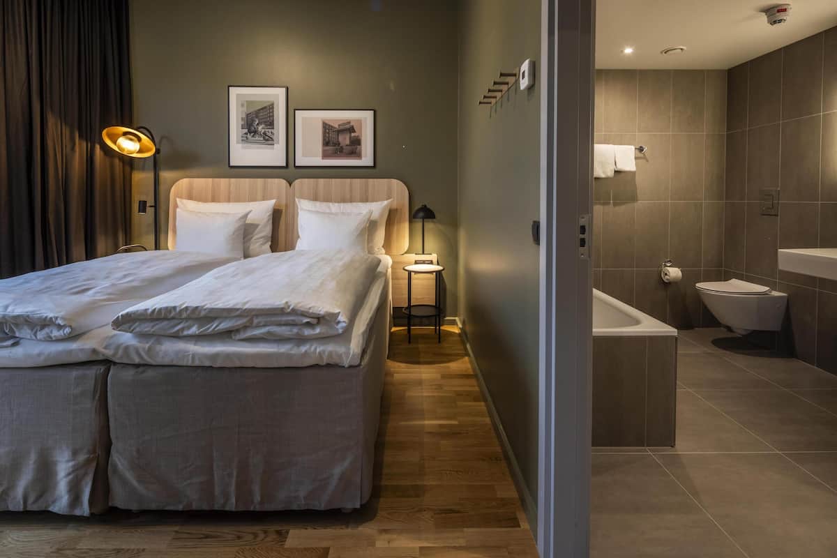 Moderni hotellihuone, jossa on suuri sänky, yöpöytä, lamppu, kehystetty taulu sekä kylpyhuone, jossa on kylpyamme ja wc.