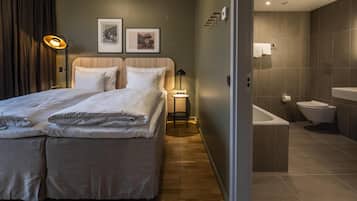 A modern hotel room with a large bed, a bedside table, a lamp, a framed picture, and a bathroom with a bathtub and toilet.