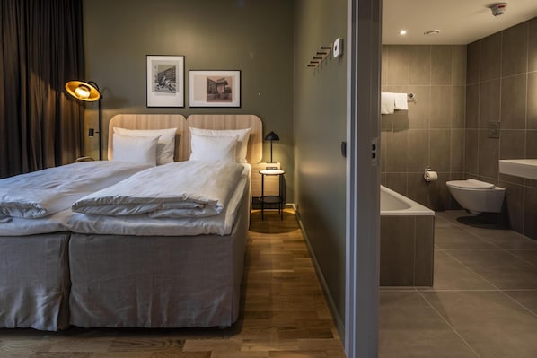 Una habitación de hotel moderna con una cama grande, una mesita de noche, una lámpara, un cuadro enmarco y un baño con bañera e inodoro.