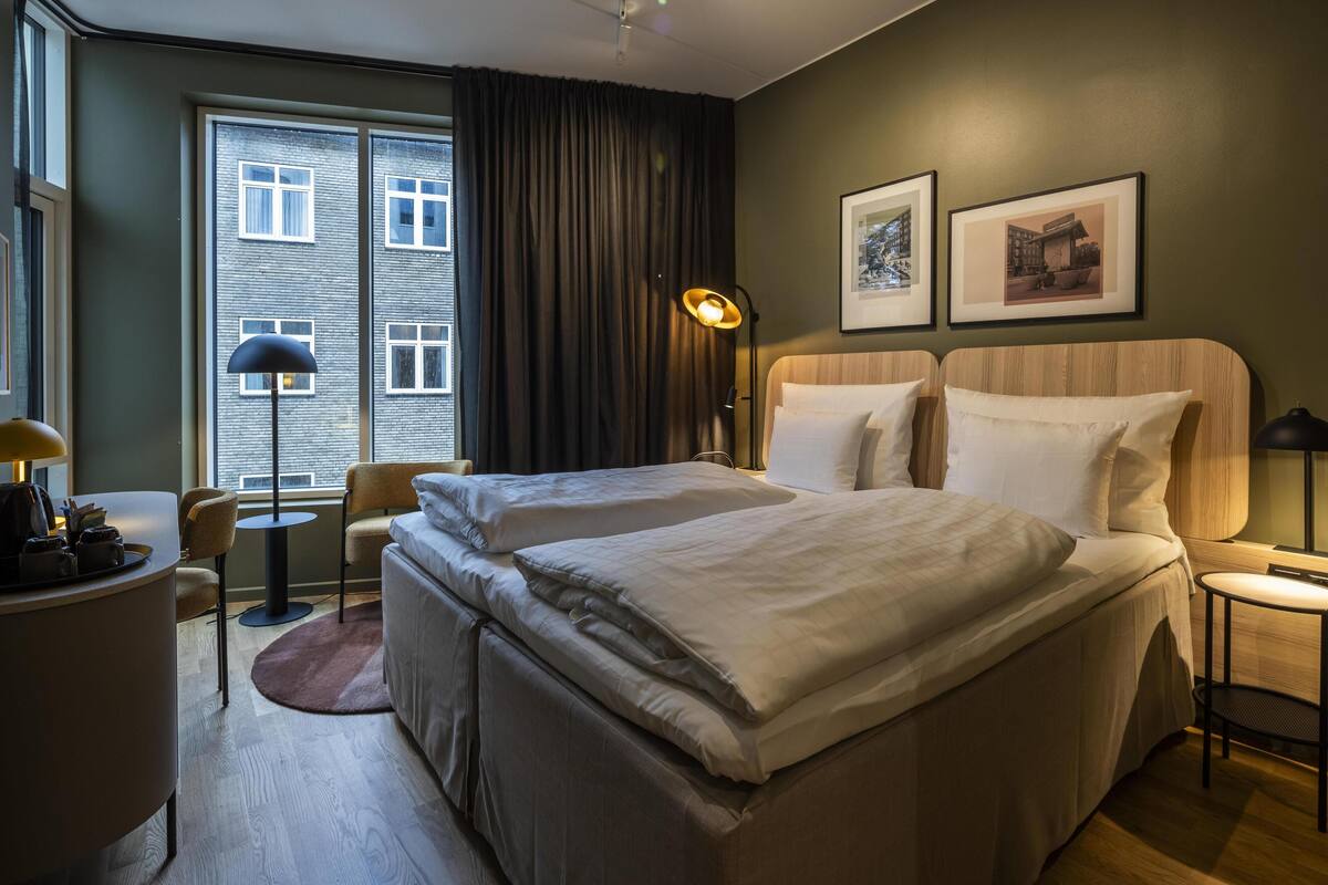 Hotellihuone, jossa on kaksi sänkyä, työpöytä, tuoli, lamppu ja ikkuna, jossa on verhot.