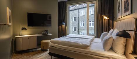 A modern hotel room with a large bed, a flat-screen TV, a desk with a lamp, and a window with a view of a building.