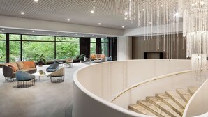En rummelig lobby med en buet trappe, moderne siddepladser og store vinduer der giver udsigt til grønne områder.