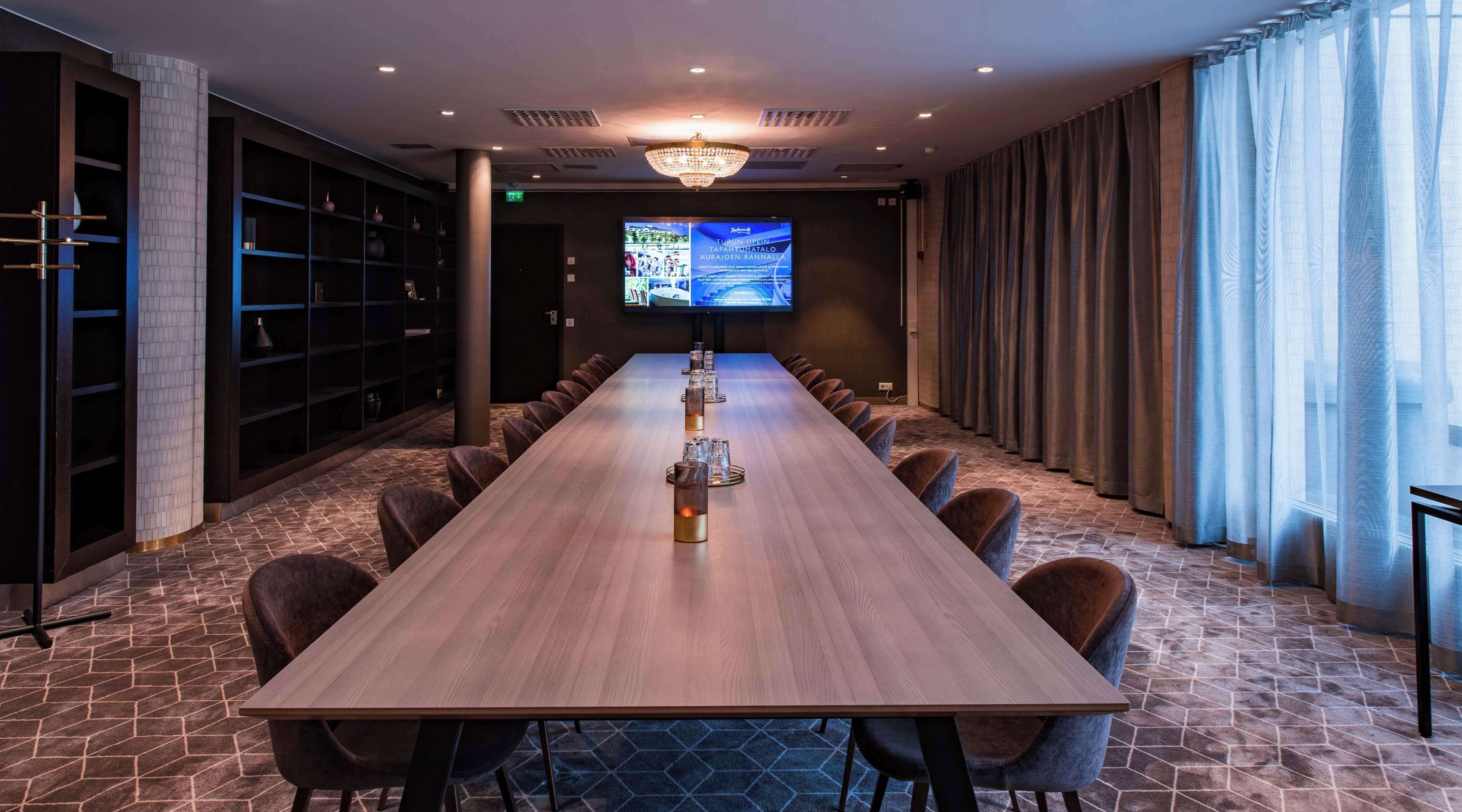 A conference room with a long table, chairs, a chandelier, a television screen, and a bookshelf.