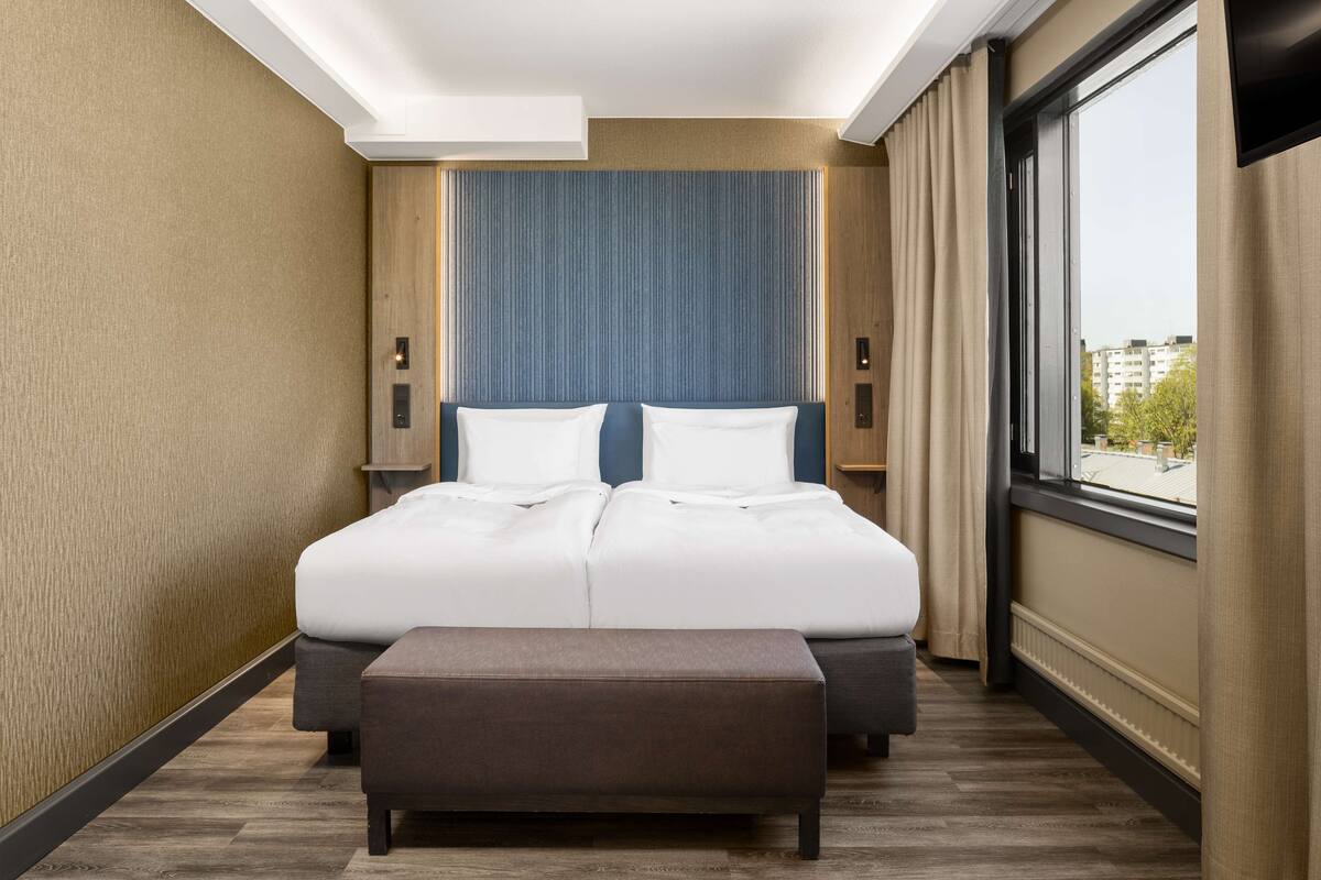 Ein Hotelzimmer mit einem großen Bett, einer Bank, einem Fenster mit Blick und einer Wand mit strukturierter Oberfläche.