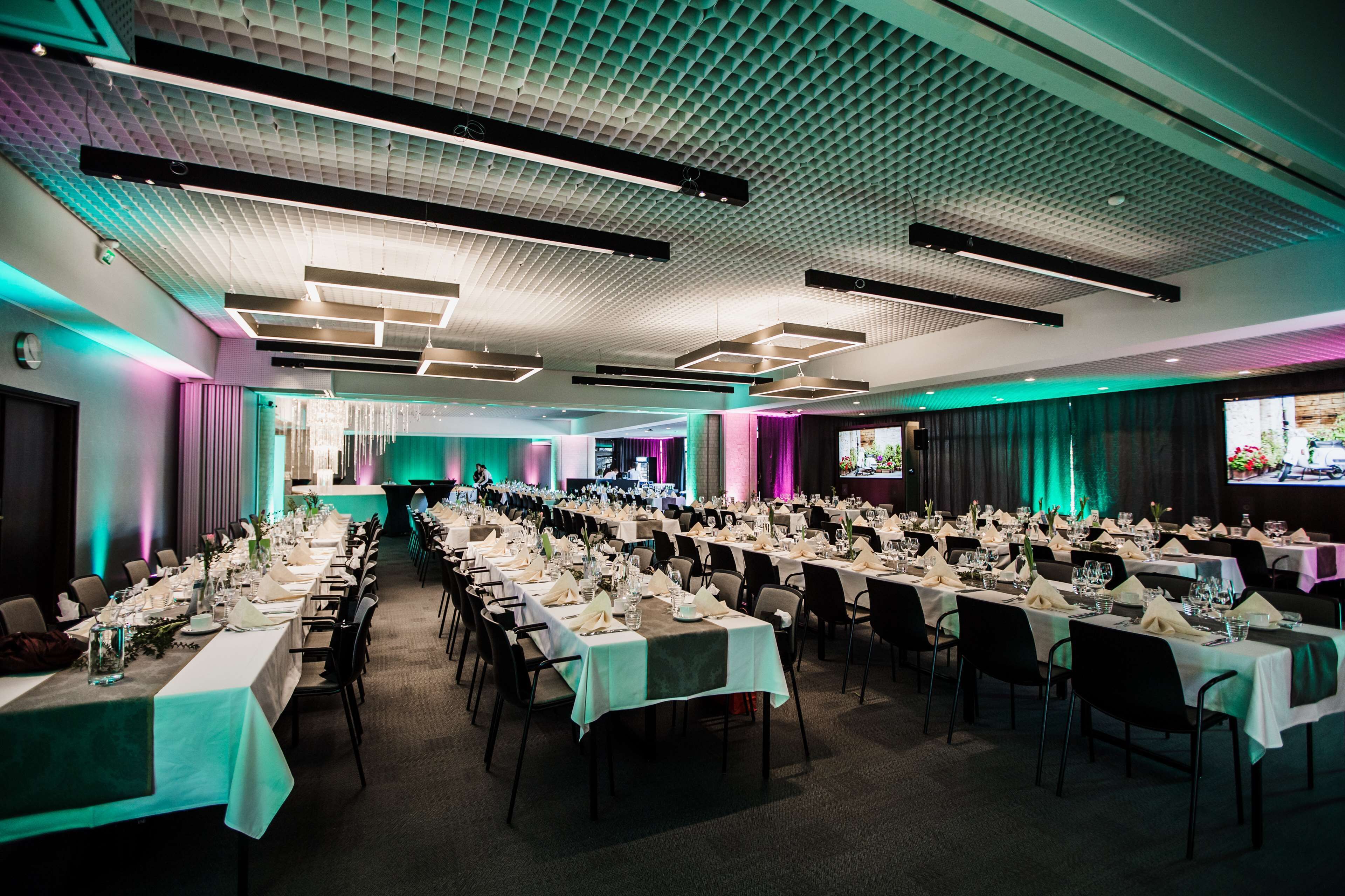 A large banquet hall with long tables set for a formal event, featuring a modern ceiling design and ambient lighting.