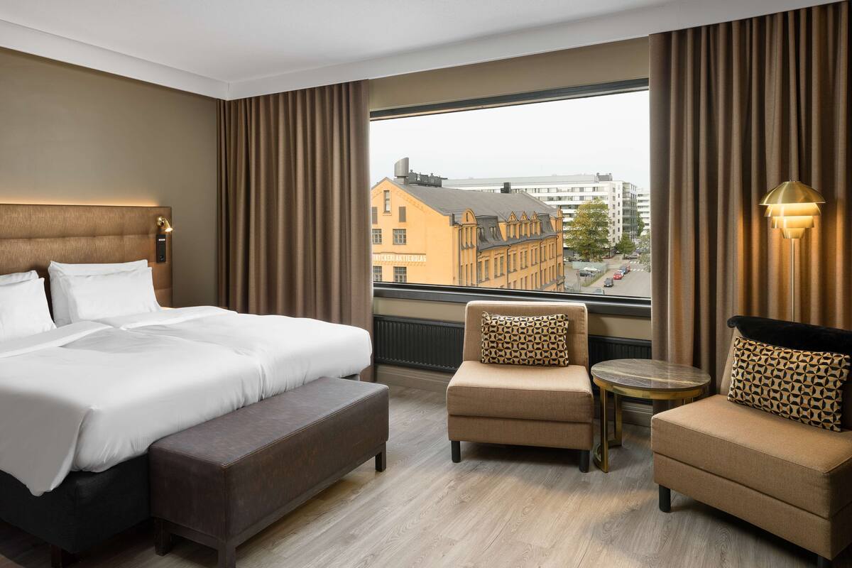 Ein Hotelzimmer mit einem großen Bett, einer kleinen Sitzecke, einem runden Tisch und Blick auf Gebäude durch das Fenster.