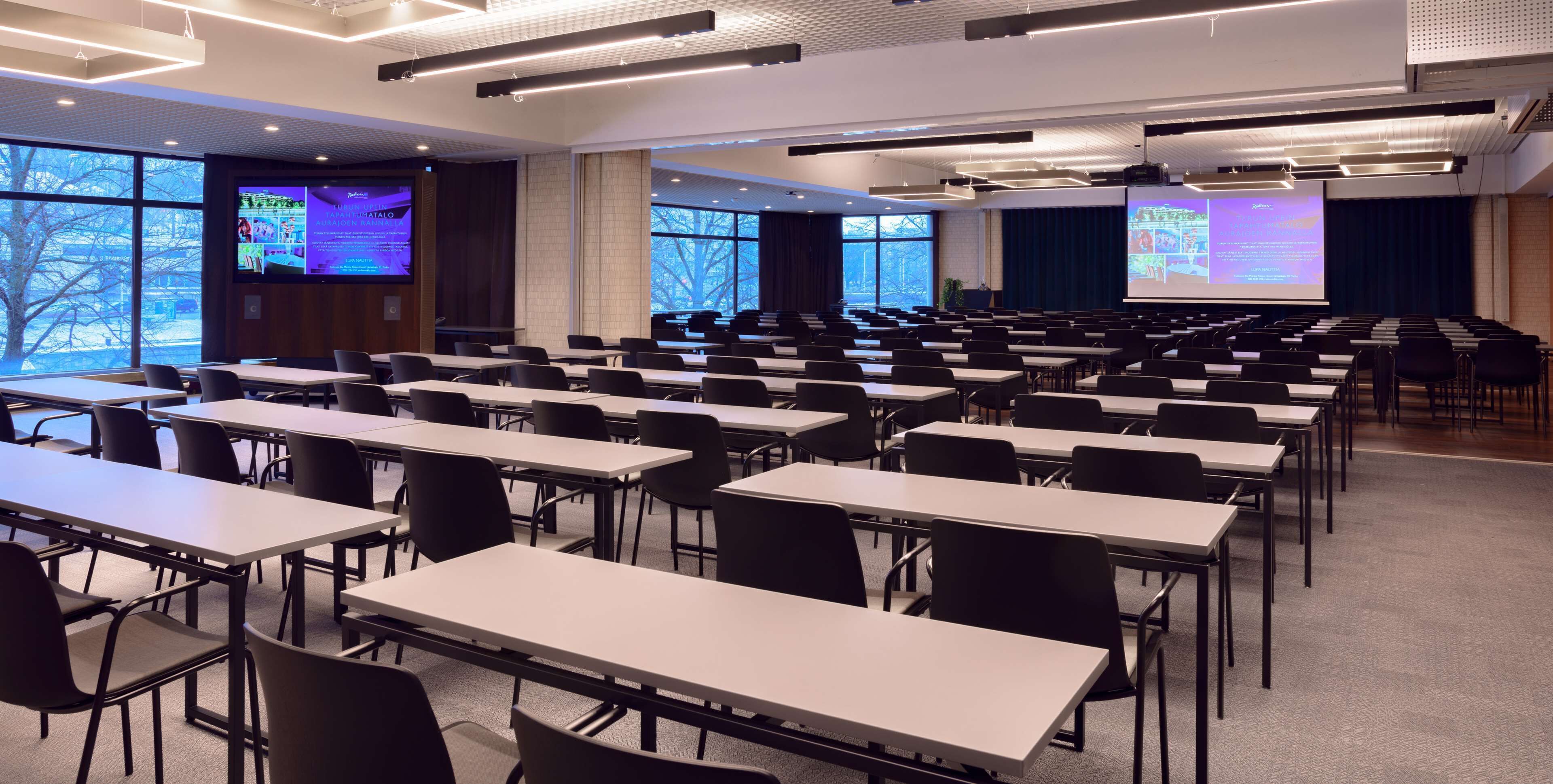 Un salón de conferencias con mesas y sillas dispuestas en filas, amplias ventanas y una pantalla de proyección.