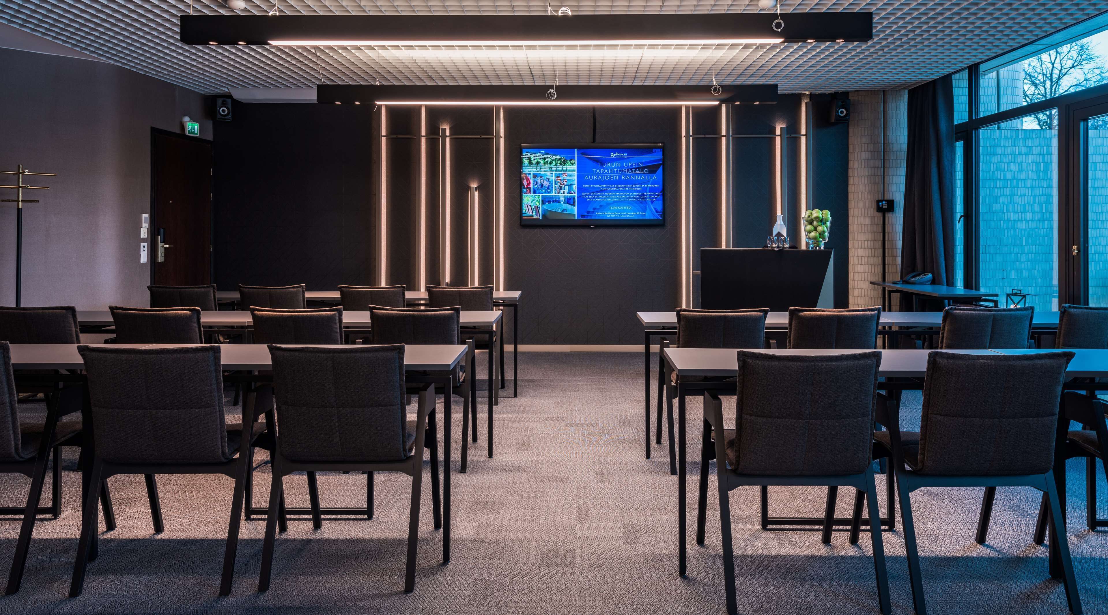 A conference room with multiple tables and chairs, a large screen displaying content, and a view of the outside through a window.